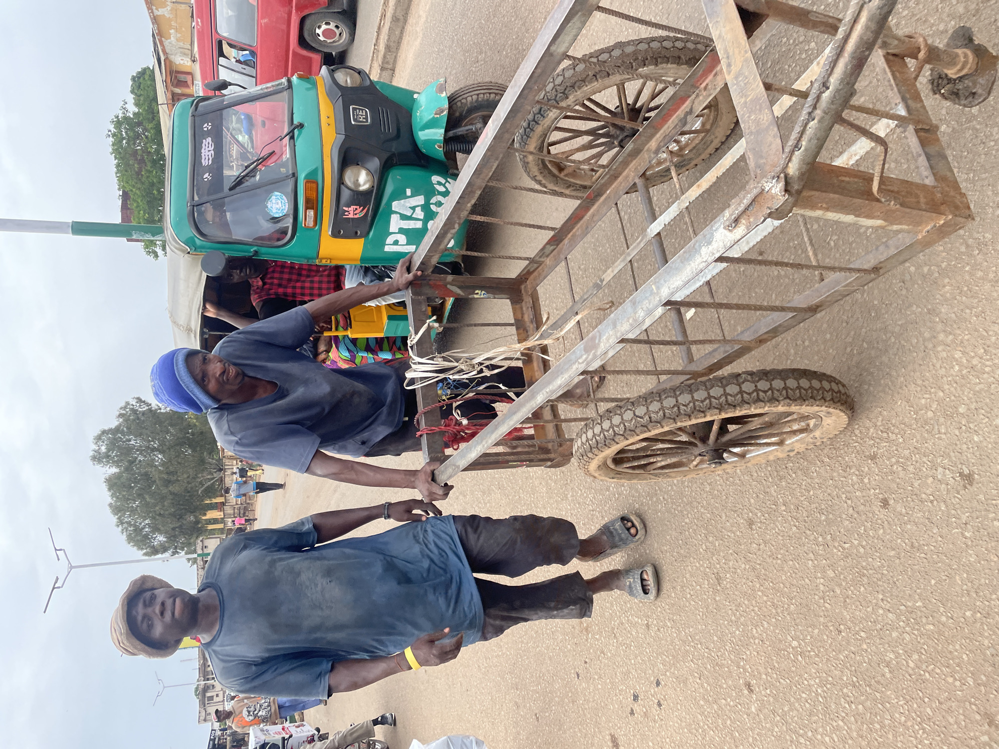 Two men are pulling a metal cart on a paved street in Jos, Nigeria. One man, wearing a blue cap and blue shirt, pulls the cart while positioned between the cart and a motorized tricycle taxi. The other man, wearing a tan cap and blue shirt, pulls from the front of the cart. The cart has two wheels and a metal frame. In the background, there is a green and yellow tricycle taxi and a red vehicle parked behind it. Additional people, buildings, and street lights can be seen in the background, suggesting an urban environment.