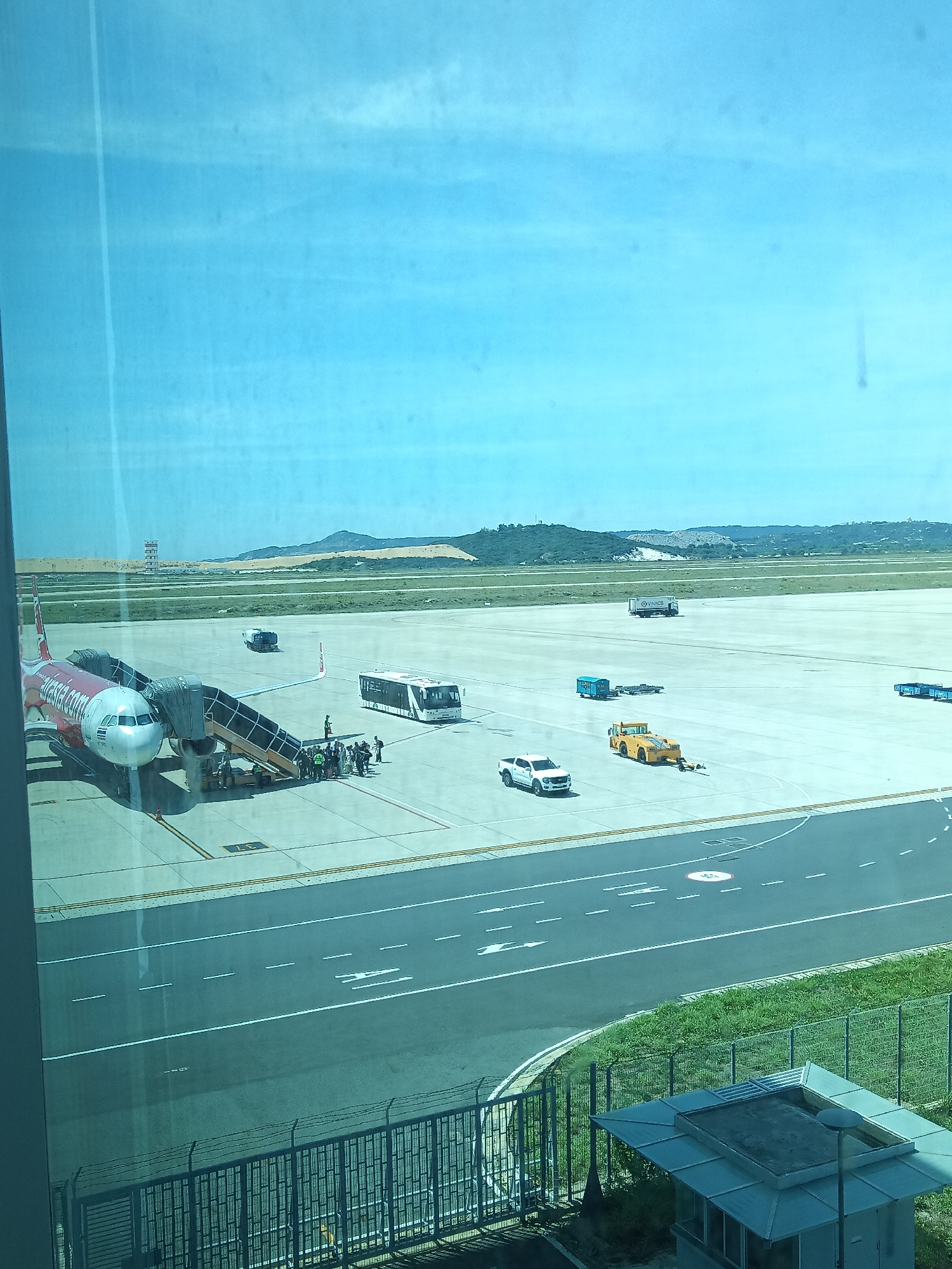 The image depicts an outdoor scene at Cam Ranh International Airport in Bắc Cam Ranh, Vietnam. The main subject is a red and white AirAsia airplane parked on the tarmac, with a mobile passenger staircase connected to its doors. Several individuals, likely passengers and airport personnel, are visible near the staircase and boarding the aircraft. In the background, a white airport bus and various ground support vehicles, including a yellow tow tractor and a white pickup truck, are present on the concrete apron. Beyond the tarmac, grassy areas and a road are visible, with a chain-link fence in the foreground. In the distance, a control tower and a landscape of hills with some construction or debris are discernible under a clear blue sky. The scene appears to capture a typical moment of passenger boarding or disembarking at an airport.
