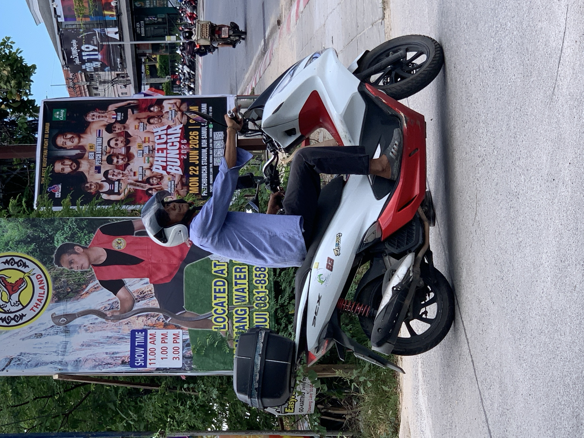 A man wearing a blue shirt, black pants, and gray sandals rides a white and red scooter in front of a poster advertising a Muay Thai fight. The scooter has a black storage box mounted on the rear. The poster on the left shows a man holding a snake with text "SHOW TIME" followed by times. The poster to the right features fighters with the text "PHETCH BUNCHA 2026". The scene appears to be on a street in Ban Rak, Thailand, with parked motorcycles and buildings visible in the background. The ground is asphalt with white lane markings.