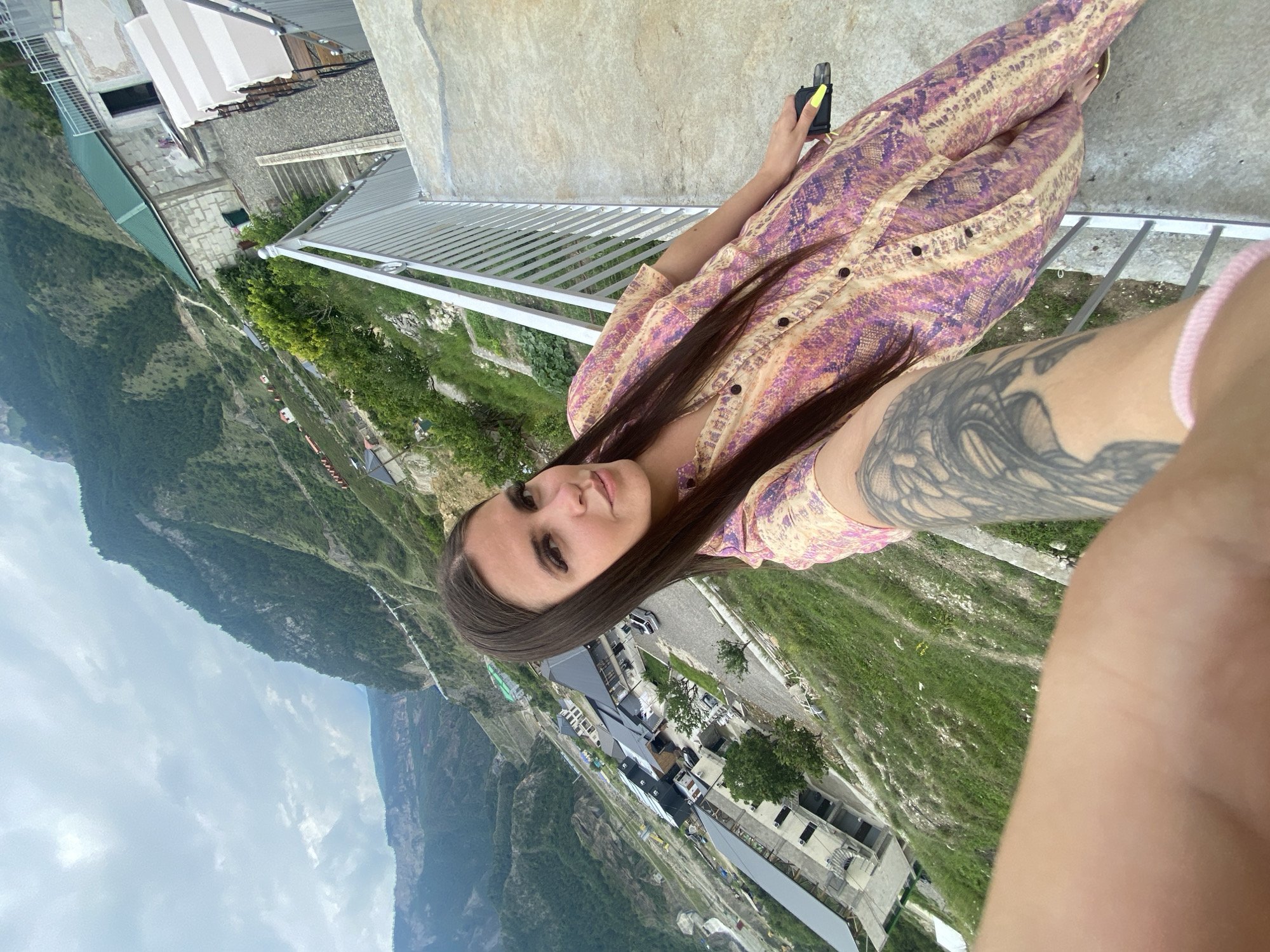 A woman with long, dark hair is taking a selfie on a concrete balcony overlooking a village nestled in a steep, mountainous valley. The village appears to be Chalda, Russia, characterized by its buildings with dark, sloped roofs and roads winding along the terrain. The woman is wearing a colorful, patterned dress and has a tattoo on her left arm. She is holding a phone in her right hand, and her left hand rests on a metal railing. The sky is overcast with clouds. The buildings in the village range from multi-story structures to smaller houses, with greenery and trees interspersed throughout the landscape. The overall scene depicts a scenic, remote location with a focus on the woman and the expansive mountain views.