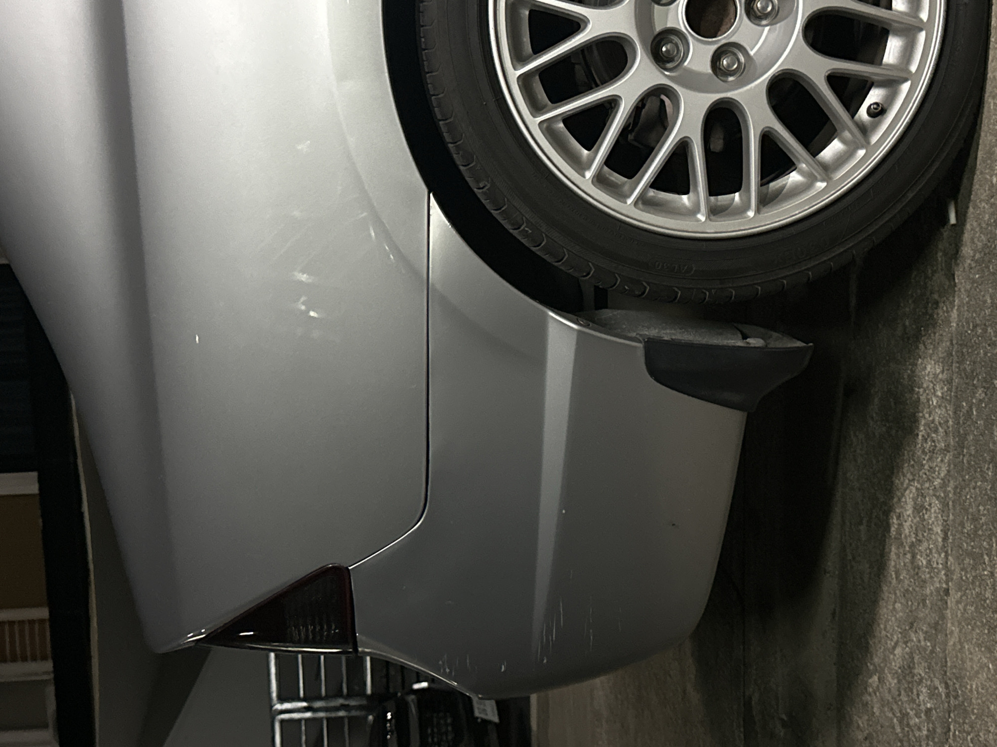A silver sedan is parked on a concrete surface. The rear passenger side of the vehicle is visible, showing the taillight, bumper, and wheel. The car exhibits several scratches on the bumper. The background suggests an urban environment with buildings and what appears to be a parking garage. The lighting is dim, indicating nighttime or an indoor setting. The image was taken in Kathu, Thailand.