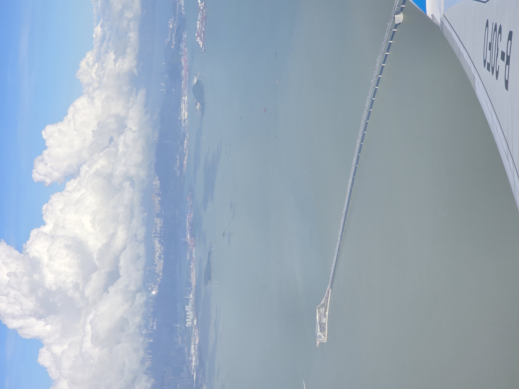 An aerial view captures the vast expanse of the Pearl River Delta, with the HZMB bridge stretching across the calm, grayish-green water. The bridge, a prominent engineering feat, connects to an artificial island which is surrounded by a breakwater. In the distance, a sprawling cityscape with numerous buildings is visible under a bright blue sky dotted with large, fluffy white clouds. Several ships and smaller vessels are scattered across the water. The wing of an airplane is visible in the foreground, with partial registration markings indicating "B-3050".