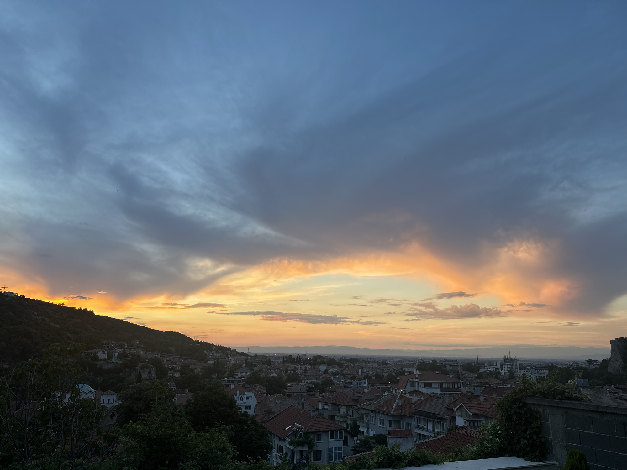 This is a wide-angle, eye-level, outdoor, landscape photograph of Asenovgrad, Bulgaria at sunset. The foreground consists of the top of a wall with some greenery. The middle ground shows a densely populated town with red-tiled roofs and white-walled buildings. The background is dominated by a mountain range and a vibrant sunset sky with clouds in hues of orange, yellow, and grey. The sun appears to be setting behind the mountain on the left side of the image. The overall atmosphere is serene and picturesque.
