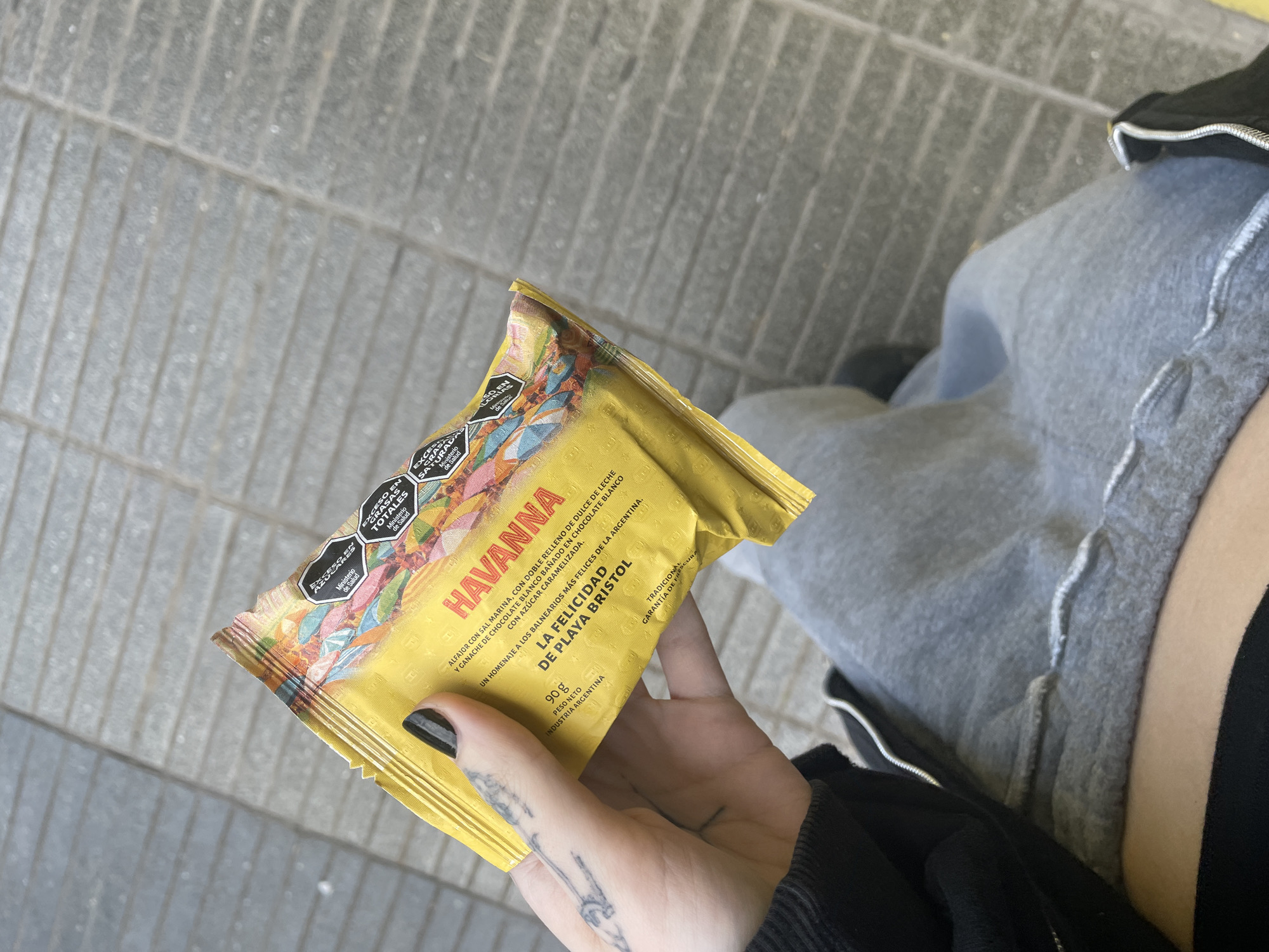 A hand holds a yellow wrapper of Havanna alfajor "La Felicidad de Playa Bristol" in front of a person seated on a grey stone or concrete surface. The alfajor wrapper is marked with "Exceso en Grasas Totales" and other health warnings. The packaging indicates it contains dulce de leche and white chocolate ganache covered in caramelized white chocolate. The person has pale skin with tattoos visible on their hand. They are wearing black nail polish and a black jacket over grey sweatpants. The packaging is from Argentina. Havanna alfajores are produced in Mar del Plata. Playa Bristol is a beach in Mar del Plata.