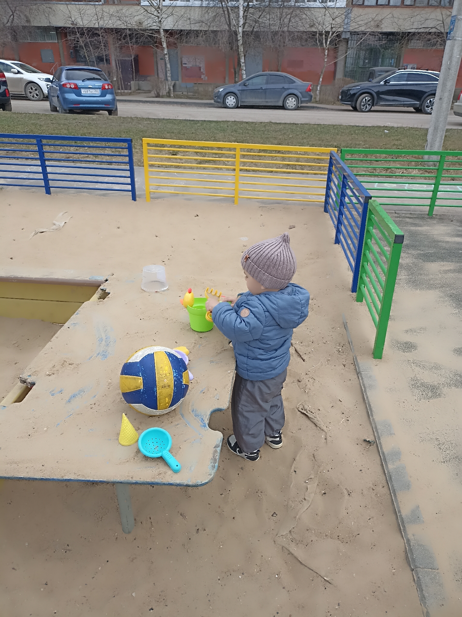 The image depicts a child playing in a sandbox in Serpukhov, Russia. The sandbox is bordered by colorful blue, yellow, and green fences. The child, wearing a blue jacket and a gray knit hat, stands at a table within the sandbox, interacting with a green bucket and various plastic toys. A blue and yellow volleyball also rests on the table. The sandbox contains loose sand, some of which covers the surfaces within. The location appears to be outdoors, with apartment buildings and parked cars visible in the background. The weather seems overcast.