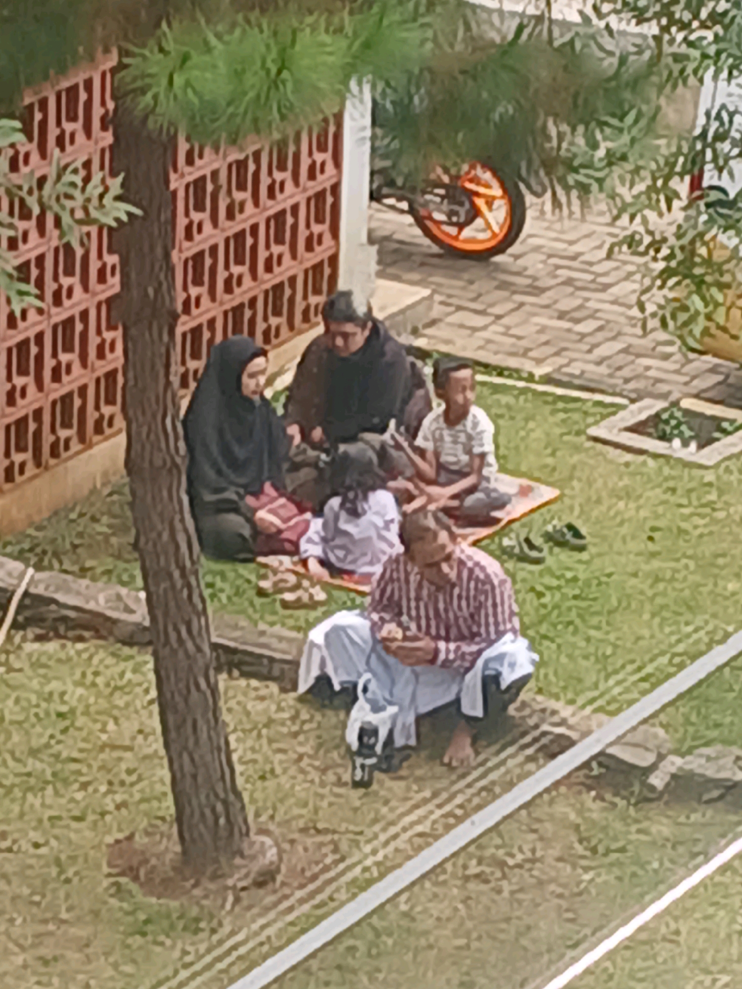 In Arjasari, Indonesia, a group of people are gathered on a grassy area outside a building with a red patterned wall. A mature tree with rough bark stands prominently in the foreground, its base surrounded by loose soil. To the right, a paved area features an orange-wheeled motorcycle.
The group consists of at least five individuals. A woman wearing a black hijab is seated on a red and white mat, facing left. Next to her, a person in a dark hooded jacket is seated. A young child in a white and black striped shirt is also seated on a mat, facing right. In front of them, a child in a light purple outfit is seated on a mat. A man in a red and white striped shirt and white pants is seated on the grass, facing left, holding something in his hands. A white cloth is draped over his lap and legs. Two pairs of dark green sandals are on the grass near the man and the child in the striped shirt. A black electronic device with a handle is positioned near the man's feet. The environment appears to be a residential or communal outdoor space, possibly a yard or garden, with a pathway or edge marked by white lines.