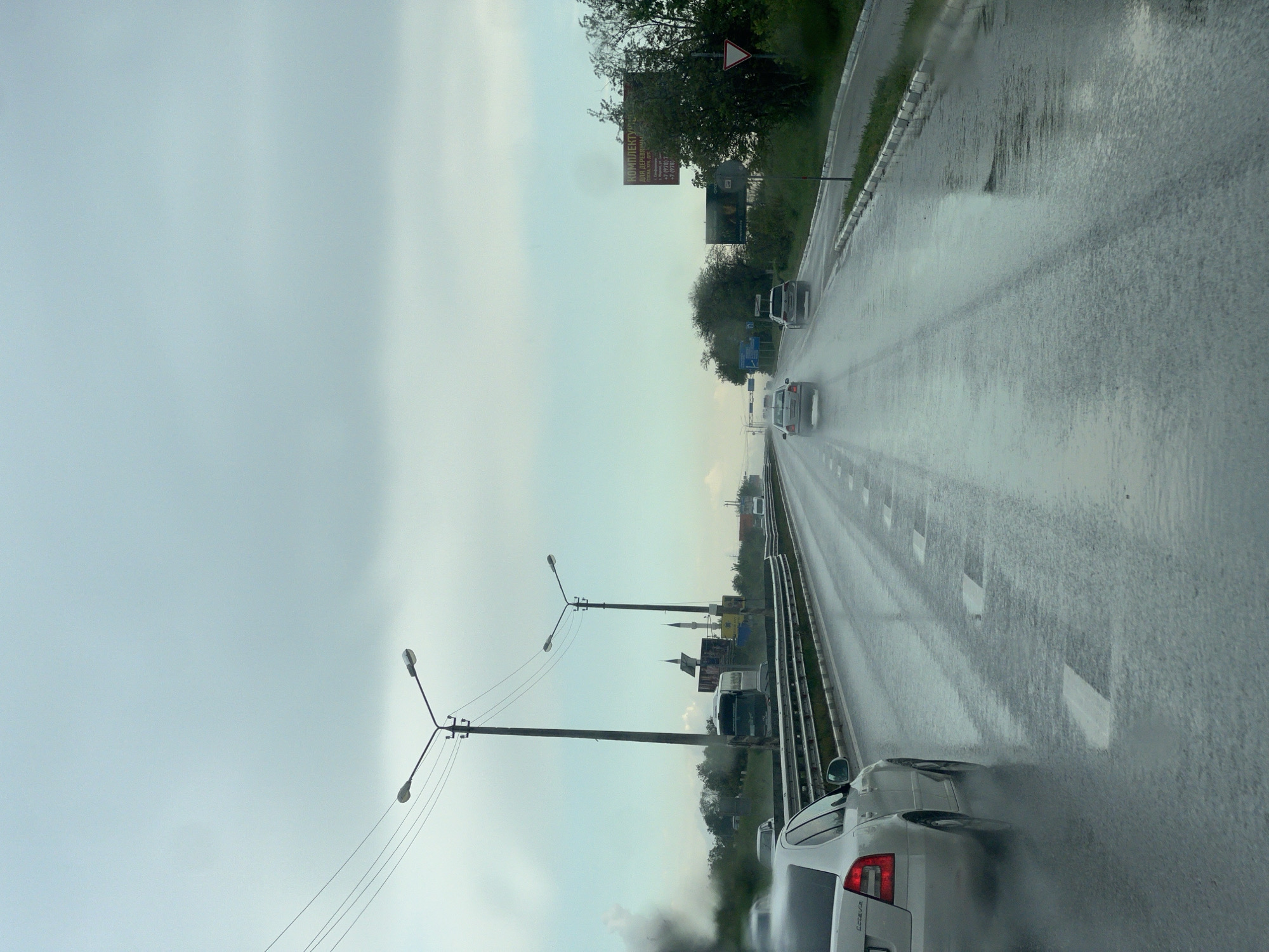 The image shows a highway scene in the city of Чистенское сельское поселение, Ukraine, during a rainy day. The road surface is wet and reflective, indicating recent or ongoing rainfall. Multiple vehicles are visible on the road, including cars and a van, moving in the same direction. Road markings are present and appear as white dashes. Lampposts are visible on the side of the road, and some signage can be seen alongside the highway. The sky is overcast, suggesting a cloudy or stormy weather. The overall impression is one of driving on a wet highway under gray skies.