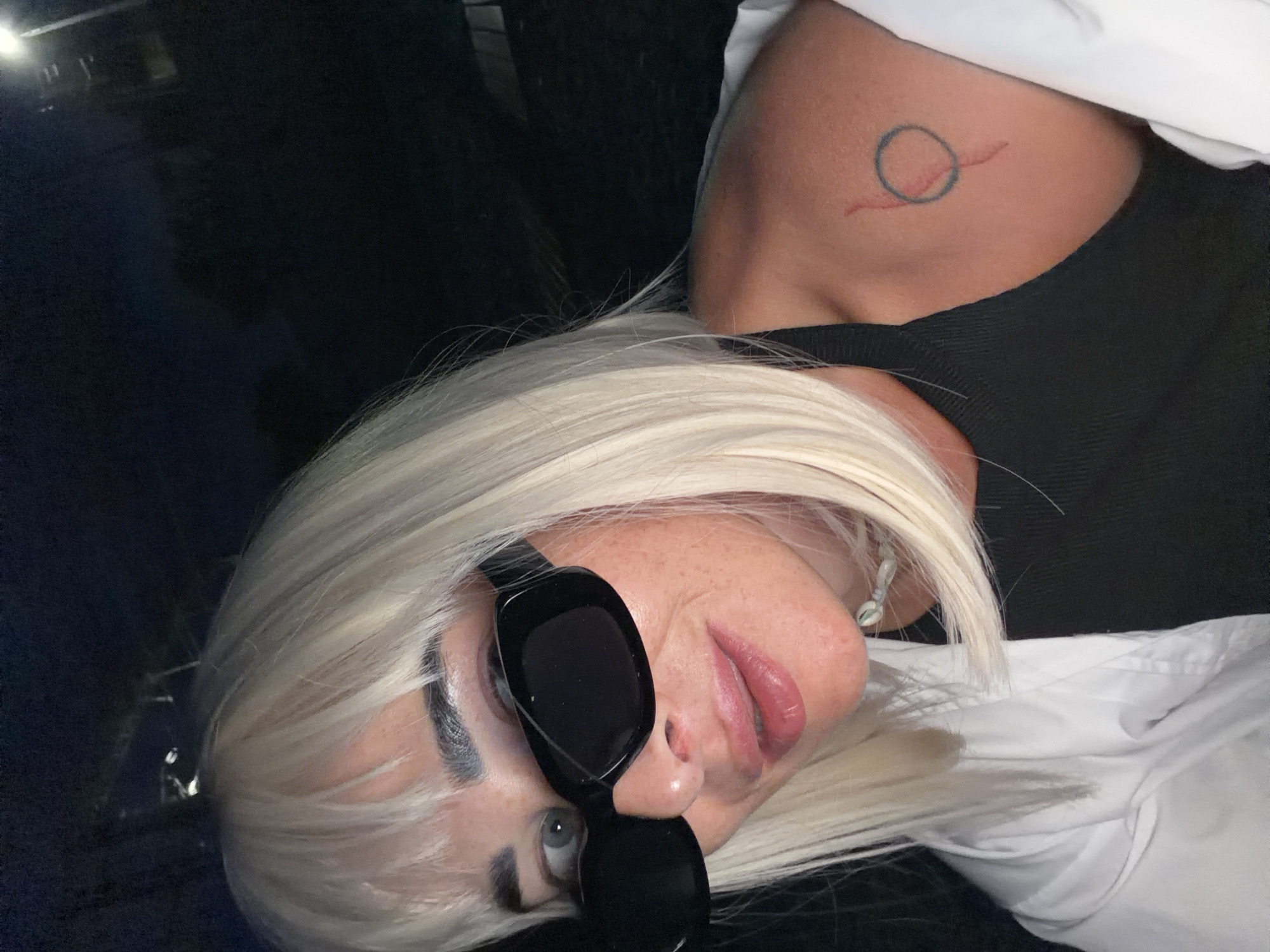 The image shows a close-up selfie of a woman with blonde hair and bangs, wearing black sunglasses and a white shirt over a black top. She has a tattoo on her right shoulder. The background is dark and out of focus, suggesting it was taken at night, possibly outdoors. There are no discernible elements within the image that can be used to identify the location as Sochi, Russia.
