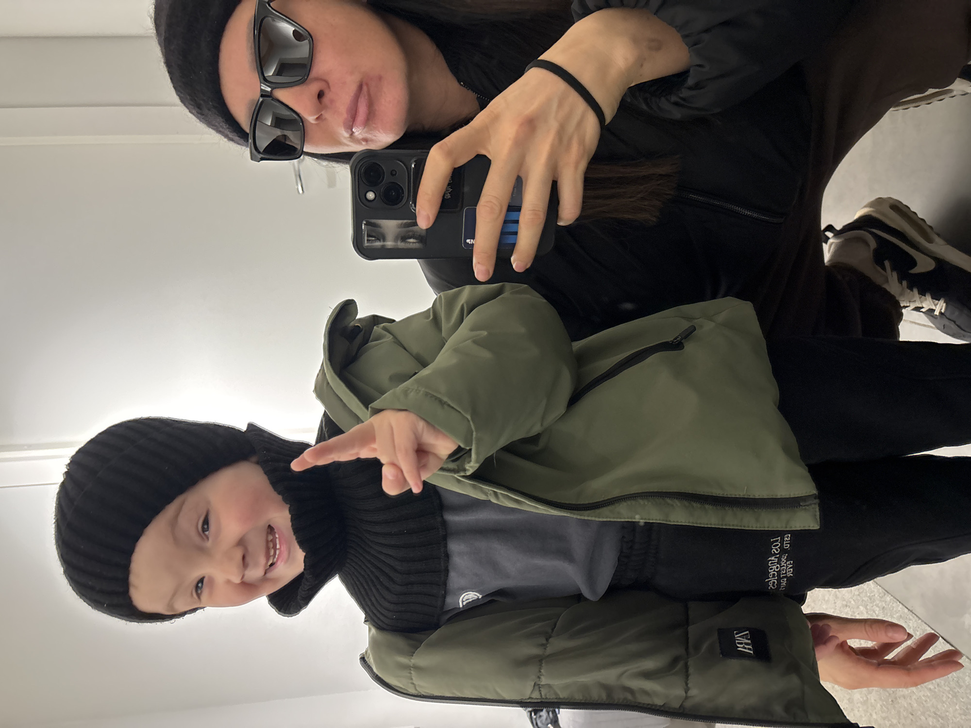 A selfie taken in what appears to be a fitting room. The adult in the upper portion of the frame wears a black beanie and sunglasses and holds a black smartphone. A younger child in the lower portion of the frame wears a black beanie, a gray shirt, and a green jacket. The child is pointing to the right. The background is a plain white wall. The brand "S&H" is visible on the child's clothing. The location, Chaikovsky, Russia, is not discernible from the image.