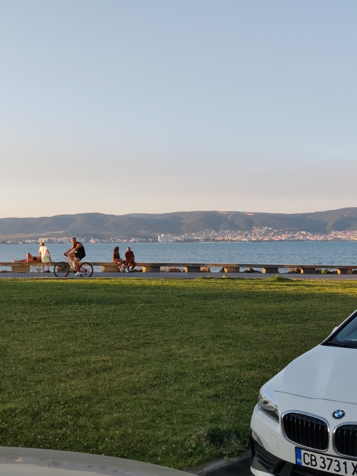 The image captures a seaside promenade in Nesebar, Bulgaria, during what appears to be late afternoon or early evening, given the soft, warm lighting. In the foreground, a patch of lush green grass is visible, dotted with small white flowers. To the right, the front corner of a white BMW sedan is parked, its license plate reading "CB 3731 X".
Beyond the grass, a paved walkway runs parallel to a low, concrete barrier that separates the walkway from the sea. On the walkway, several individuals are present. A man on a bicycle is in motion, heading towards the left of the frame. To his left, a woman reclines on the concrete barrier, appearing to sunbathe. Further along the barrier, two individuals sit together, facing the sea.
The middle ground is dominated by the clear, blue sea, with a gentle ripple on its surface. In the background, across the water, lies the city of Nesebar, its buildings clustered on a hilly peninsula, illuminated by the warm sunlight. The horizon is marked by a range of low, shadowed hills. The sky above is a clear, pale blue, transitioning to a lighter hue near the horizon, suggesting the approaching sunset.