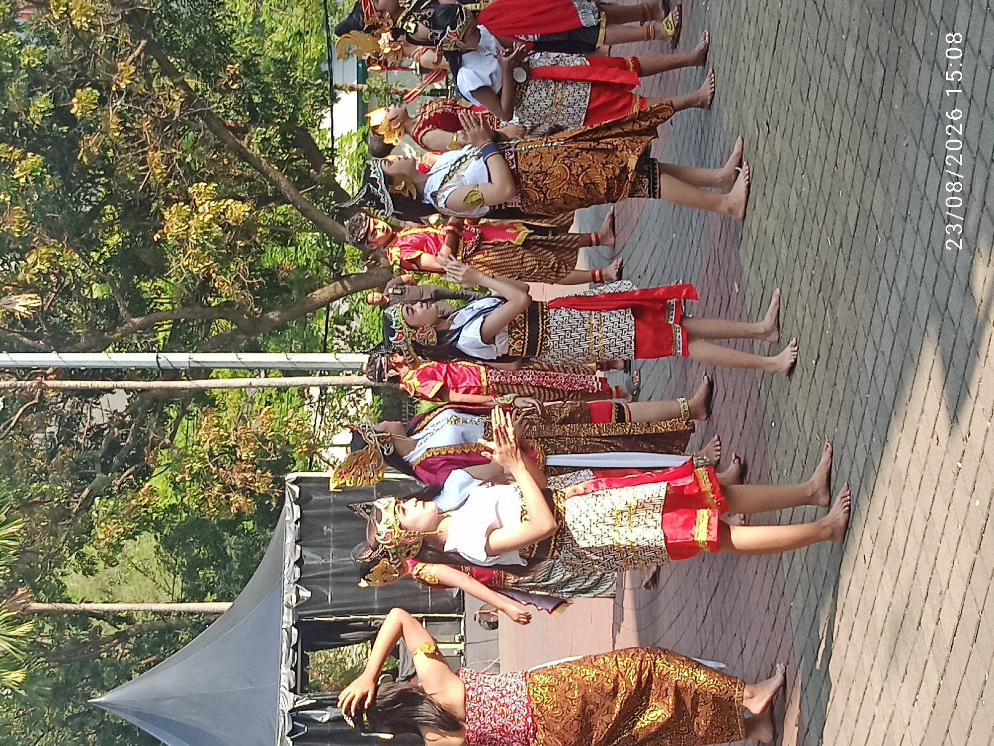A group of young people, likely performers, are dressed in traditional Indonesian attire, possibly for a cultural event or performance. They are standing on a paved outdoor area with trees and a black tent in the background. Several individuals are wearing elaborate headdresses, ornate tops, and patterned sarongs or skirts. Some are looking towards the right of the frame, while others are engaged in movements, with one person in the foreground clapping their hands. The scene appears to be set in an open space suitable for public gatherings, with dappled sunlight suggesting it is daytime. The timestamp "23/08/2026 15:08" is visible in the lower right corner. The attire and headdresses suggest a Javanese or Balinese influence. The location is specified as Semarang, Indonesia.