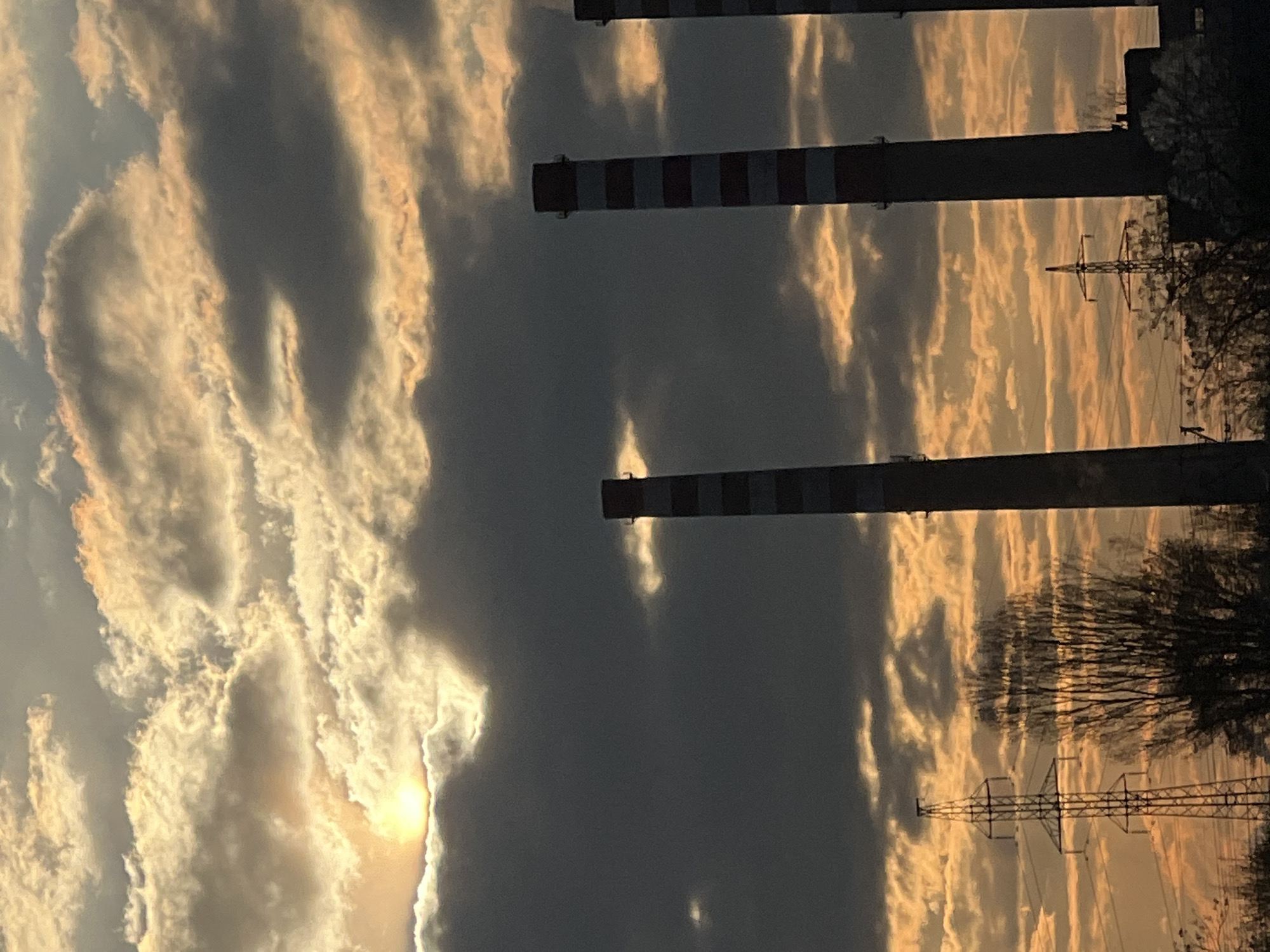 The image shows several tall, striped chimneys against a dramatic sky with dark storm clouds and patches of bright, golden light, likely at sunset or sunrise. In the foreground, a few electrical pylons are visible, along with silhouetted trees. The scene is located in Sofia, Bulgaria.