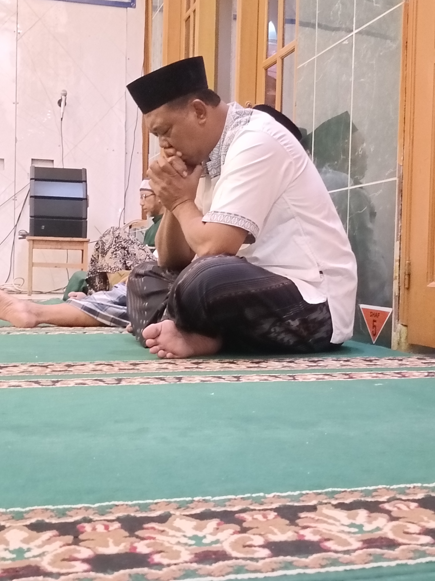 The image shows a man wearing a black peci and a white shirt with patterned trim, sitting on a green carpet in what appears to be a mosque or prayer hall. His hands are clasped in front of his mouth, suggesting he is praying or in deep thought. Another person in Islamic attire is visible in the background. The environment includes a wall with what looks like a tiled surface, a wooden door, and some electronic equipment on a small table. A triangular sign with the word "SHAF" and the number "5" is affixed to the wall. The carpet has decorative borders. This scene likely depicts a prayer gathering or personal devotion.