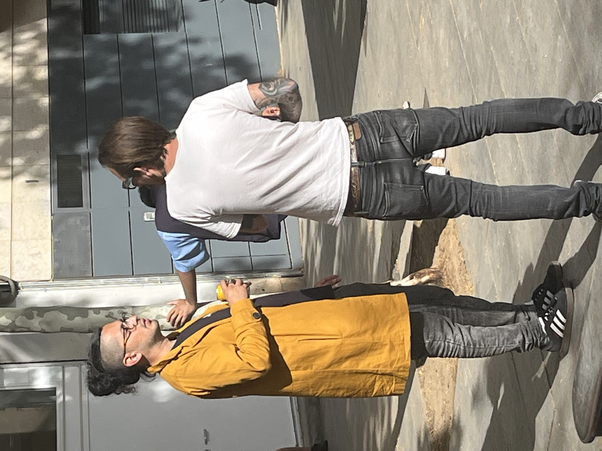 This image, taken in Barcelona, Spain, depicts an outdoor scene on a sunny day, with two men interacting on a paved surface next to a building.
In the foreground, a man with dark hair, styled in an undercut with a man bun, wears glasses and has a dark beard. He is dressed in a mustard yellow long-sleeved jacket, dark pants, and black Adidas sneakers with white stripes. He holds a yellow can in his right hand and is looking upwards and to his right, appearing to be speaking or reacting to something. A dark strap, possibly from a bag, crosses his chest.
Behind him, slightly to the right, stands another man with light brown hair, visible tattoos covering his right arm and shoulder. He wears a white short-sleeved t-shirt, dark blue or black jeans, and a brown belt with a prominent buckle. He is positioned with his back mostly towards the camera, looking down and to his left, seemingly engaged in conversation with the first man. Part of a third person, wearing a blue and white striped shirt, is visible behind him.
The setting appears to be an urban or semi-urban outdoor area, with a modern-looking building facade featuring large grey panels and a window or door. Strong sunlight creates sharp, dappled shadows on the ground and building, indicating a bright daytime environment. The ground is paved, likely concrete or stone. The overall activity suggests a casual social interaction between the individuals. The only visible text is the Adidas brand stripes on the sneakers of the man in the yellow jacket.