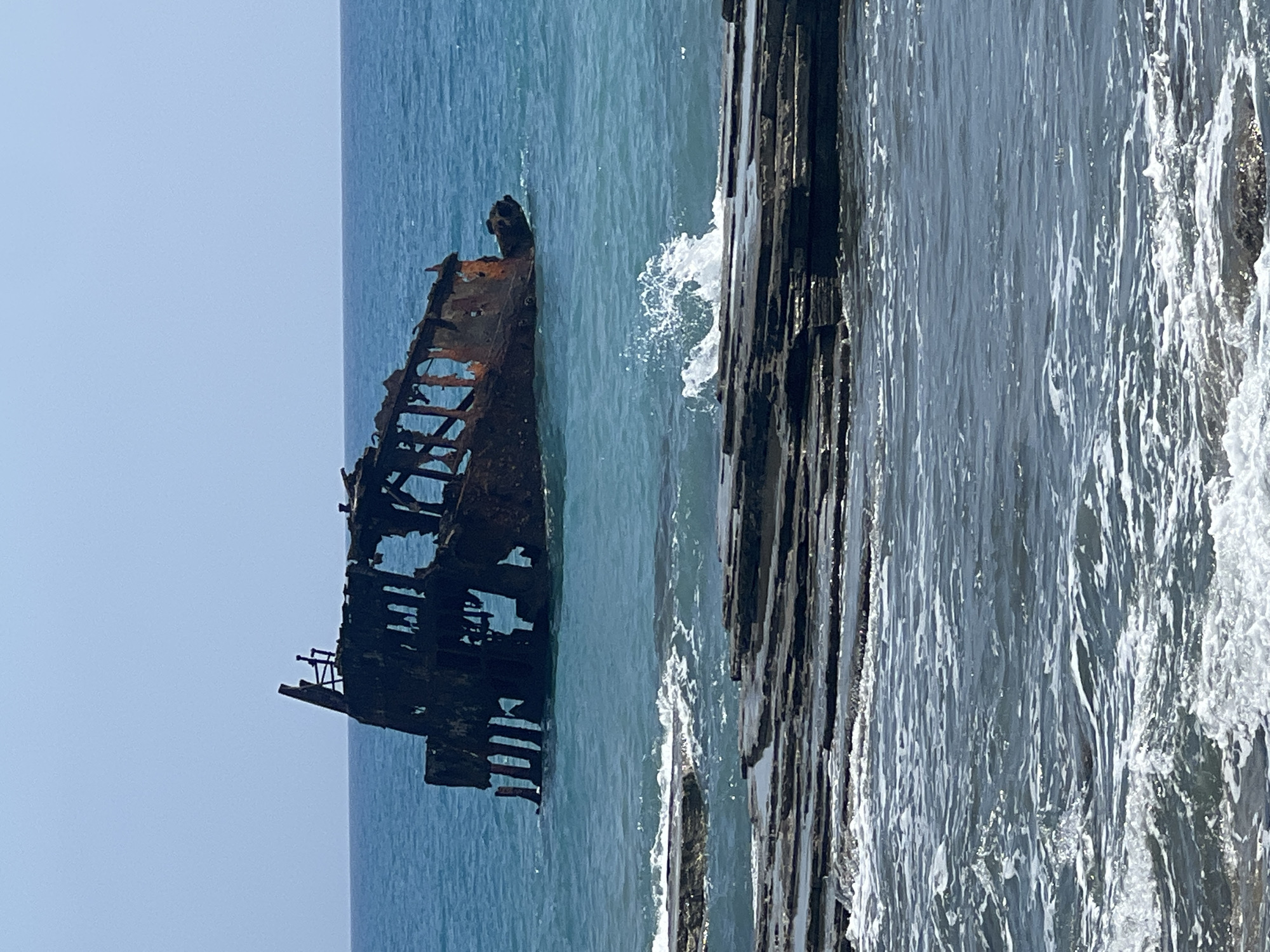 A shipwreck, rusted and broken, sits partially submerged in the blue waters off the coast of Akrotiri, Sovereign Base Areas of Akrotiri and Dhekelia. The skeletal remains of the ship, a dark silhouette against the bright sky, are weathered and decaying. Waves crash against the ship and the rocky shoreline in the foreground, creating white foam and spray. The water is a vibrant turquoise, reflecting the clear, pale blue sky. The scene captures a sense of abandonment and the power of nature reclaiming man-made structures.