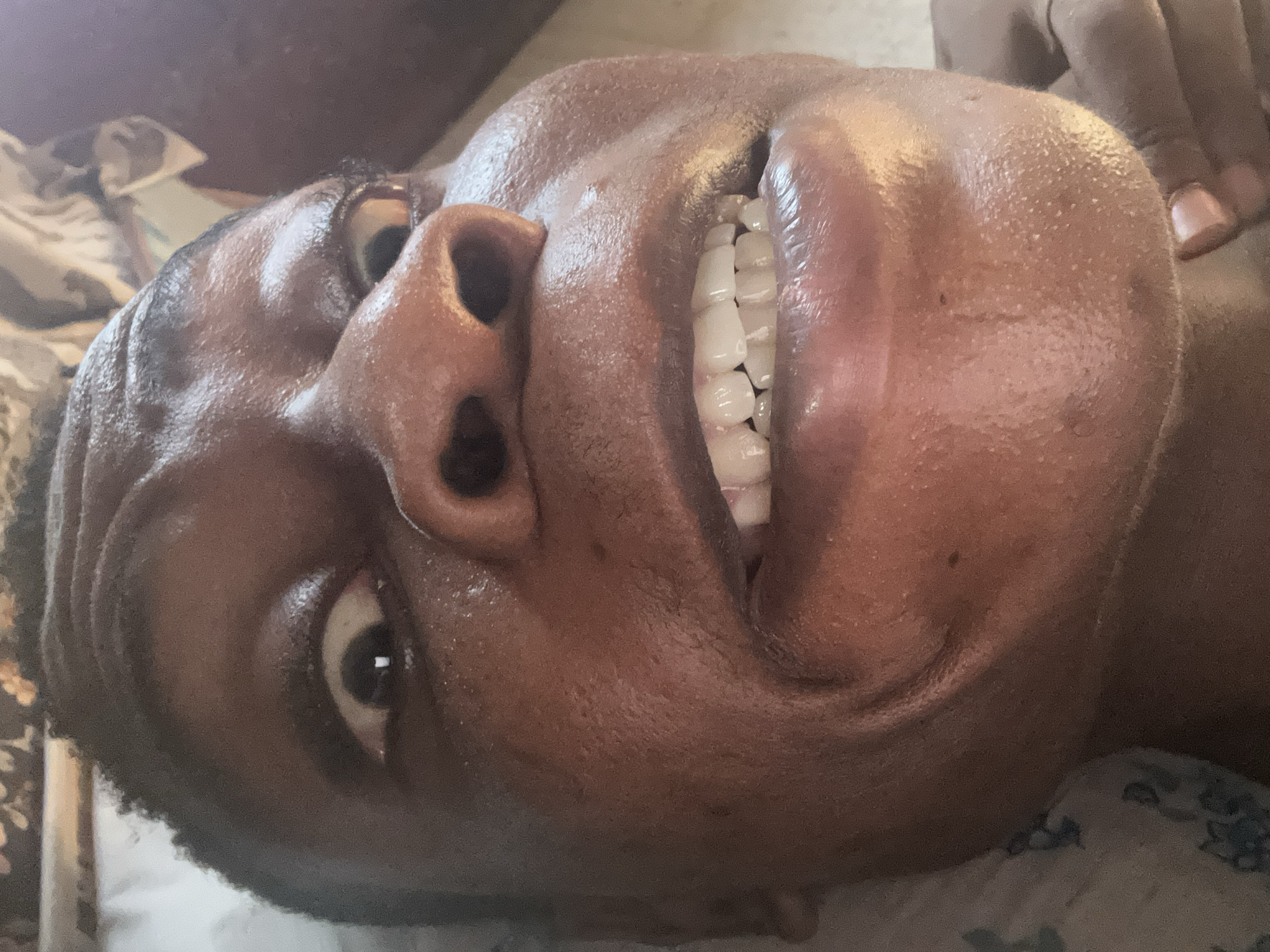 The image is a close-up, low-angle shot of a person's face. The individual has dark skin, and their face appears to be slightly moist or oily. They are smiling, revealing their teeth. The background is slightly blurred, with hints of fabric or bedding suggesting they might be lying down. The location and specific details beyond this are not discernible from the image.