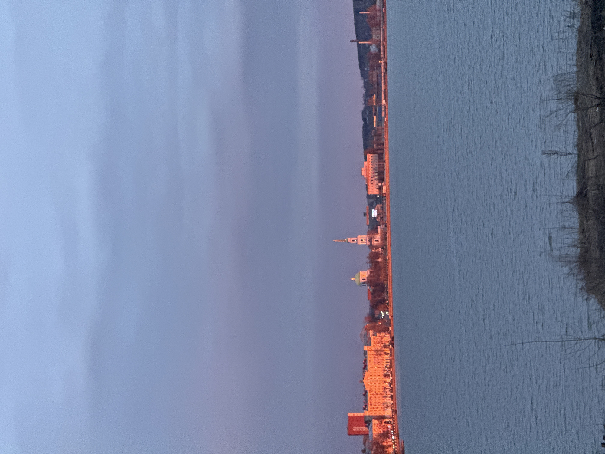 The image, correctly oriented horizontally, presents a serene panoramic view of a city skyline across a body of water during dusk or early evening. The upper left portion of the frame is dominated by a vast sky, exhibiting a gentle gradient from a light periwinkle blue to a soft purplish-pink hue near the horizon, indicating the remnants of a sunset.
Running horizontally across the middle, the cityscape features numerous buildings, many of which are illuminated with artificial lights, casting warm, orange tones on their facades. A notable landmark is a tall, slender white spire or tower, topped with a dark, possibly domed structure, rising prominently against the twilight sky. Nearby, another building is distinguishable by its bright green dome. The architecture appears to be a mix of styles, interspersed with the dark silhouettes of bare trees.
The lower right section of the image is occupied by a wide expanse of dark blue-grey water, likely the Votkinsk Reservoir, showing subtle ripples across its surface. The far bank, on the extreme right, reveals shadowy land and the intricate, bare branches of trees, suggesting a late autumn or winter season. No specific activities or people are visible, and there is no discernible text. The scene conveys a tranquil and contemplative atmosphere, capturing the calm transition from day to night over Votkinsk, Russia.