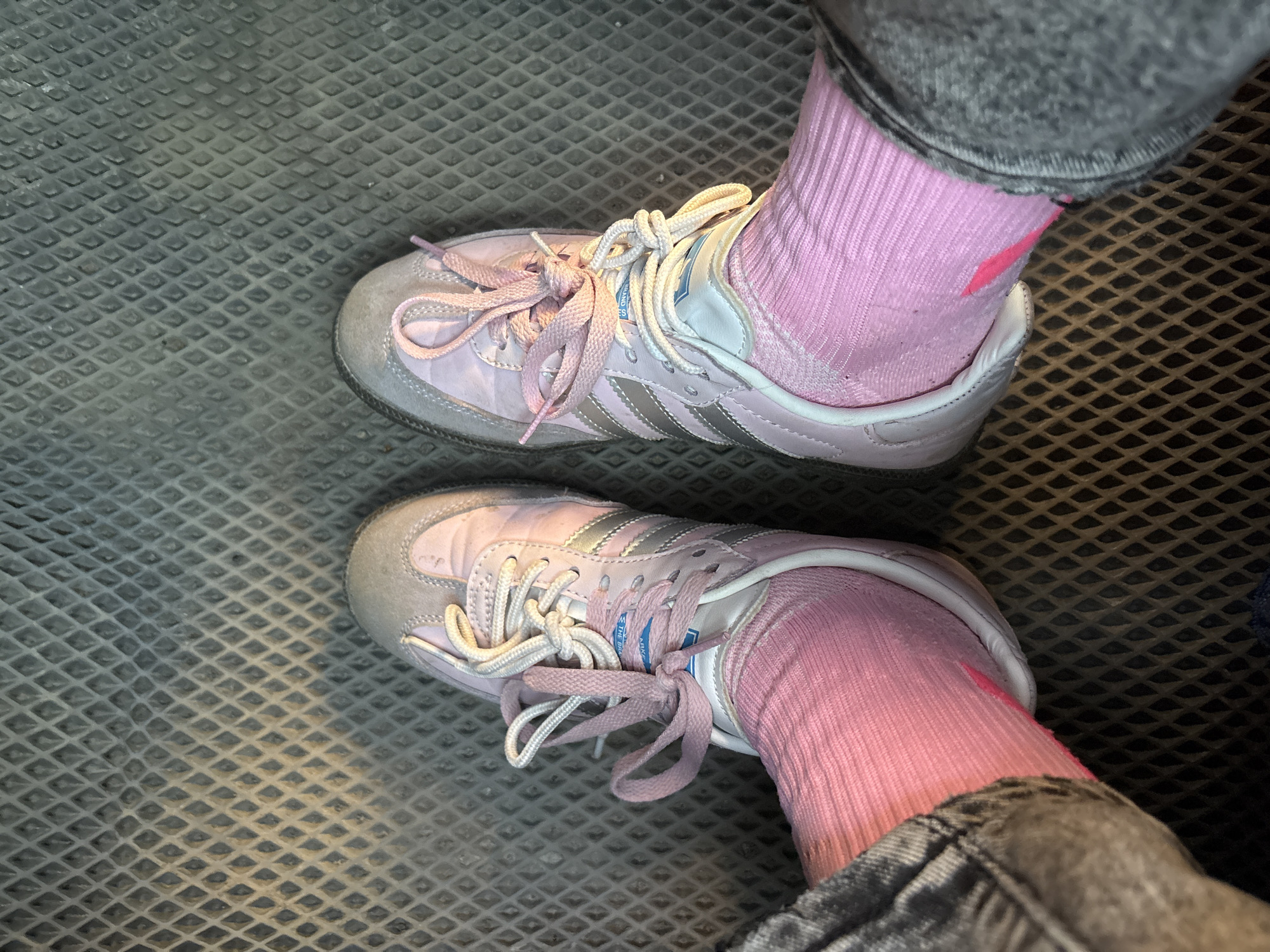 The image shows a top-down view of a person's feet wearing pink socks and pink Adidas Samba sneakers with grey suede toe caps. The person is wearing grey jeans. The footwear rests on a black, diamond-patterned metal grate surface. The location is not discernable from the image.