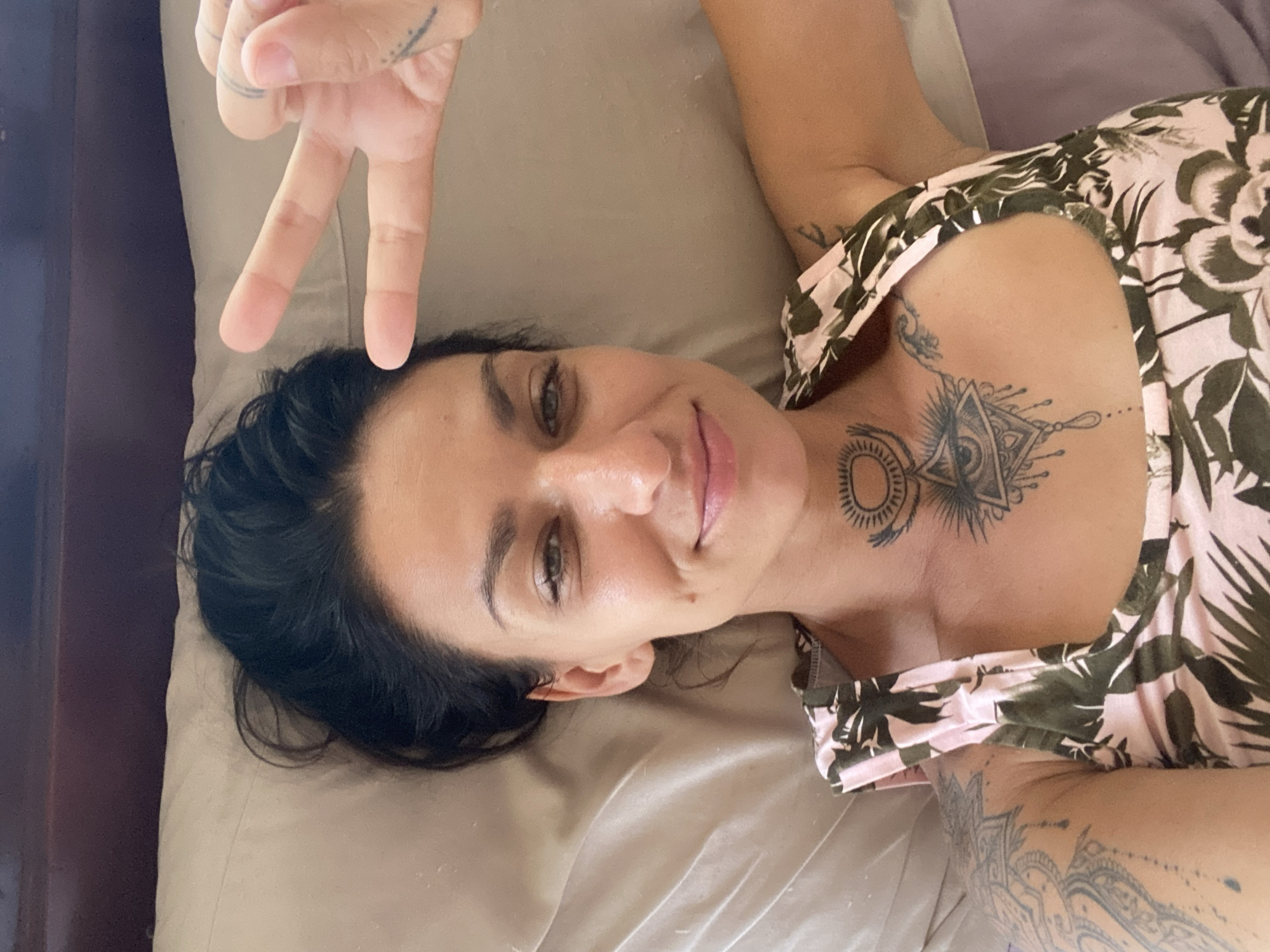 A woman is lying on a bed, propping herself up with one hand while holding up two fingers on the other hand in a peace sign. She has dark hair and is wearing a pink and green patterned tank top. She has tattoos on her chest, neck, and left arm. The background is a pillow and a headboard, suggesting an indoor setting. The scene is indoors, not in a specific city location in Mui Ne, Vietnam, as it depicts a bedroom.