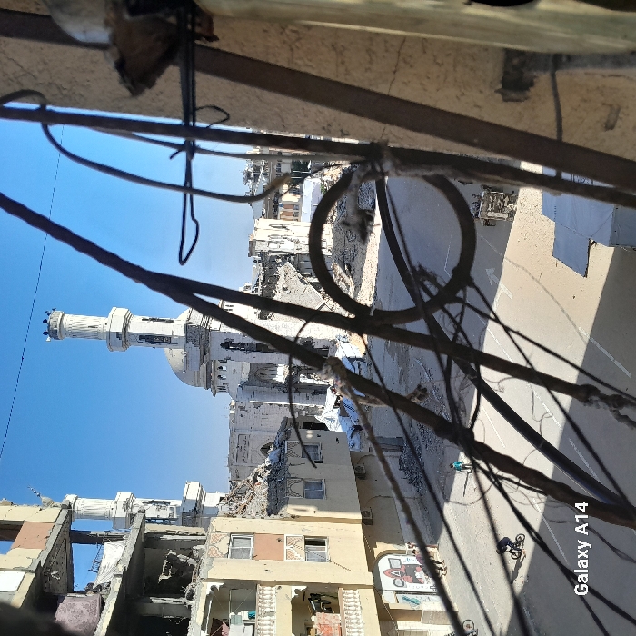 The image depicts a severely damaged urban street scene in Khan Yunis, Palestinian Territory, viewed from an elevated perspective through dark, intertwined metal bars in the foreground. A prominent mosque, painted white, stands in the mid-ground with a central dome and two minarets, exhibiting extensive structural damage, including a partially collapsed dome and visible breaches in its walls and minarets. To the left, a multi-story residential building also shows significant destruction, with its facade extensively damaged, exposing interior rooms, and large amounts of rubble accumulated at its base. Geometric artwork is visible on the ground floor exterior of this building. The paved street below appears mostly clear but shows signs of disrepair, with white directional arrows painted on its surface.
Several individuals are present on the street: a child rides a bicycle in the lower-left foreground, another child stands alone near the center of the street, and multiple adults are visible near the base of the damaged residential building on the left. In the background, further urban structures are visible, also exhibiting varying degrees of damage and collapse, with some debris piles covered by white tarpaulins. A small, damaged vehicle or piece of equipment is situated on the far right side of the street. The sky is clear and blue.