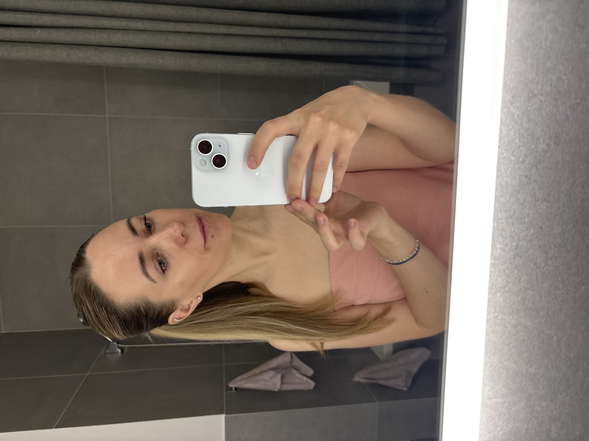 A woman with blonde hair and blue eyes is taking a selfie in a bathroom mirror. She is holding a white smartphone in her right hand. She is wearing a pink sleeveless top and a bracelet on her left wrist. The bathroom has grey tile walls, a grey shower curtain, a white mirror with an illuminated frame, and grey towels on a rack. There is no information to confirm this is located in Chaikovsky, Russia.