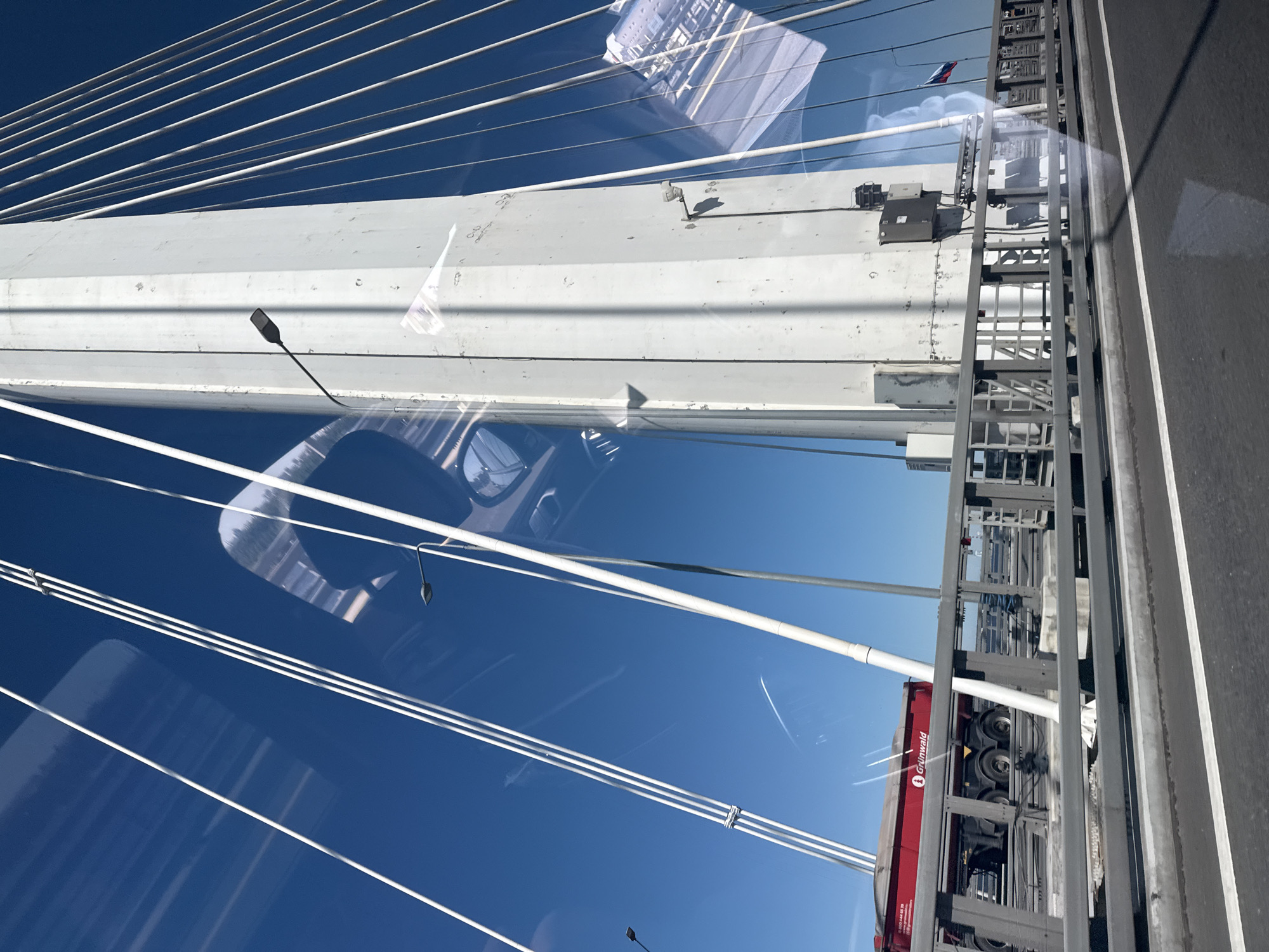 This is a medium shot, eye-level, outdoor photograph taken from a vehicle moving across a bridge. The primary subject is a large, white, pylon of a cable-stayed bridge, dominating the right side of the frame. Numerous white cables extend from the pylon upwards and outwards, creating a repeating diagonal line pattern against the clear blue sky. A red dump truck with the logo "Grünwald" is visible on the bridge deck below and to the left of the pylon. A metal railing runs along the edge of the bridge deck. Reflections of the surrounding environment, including trees and vehicles, are visible on the glass surface from which the photo was taken, superimposed over the bridge structure. A small Russian flag is visible on a pole in the distance. This bridge is the **Western High-Speed Diameter (WHSD)** in Saint Petersburg, Russia.