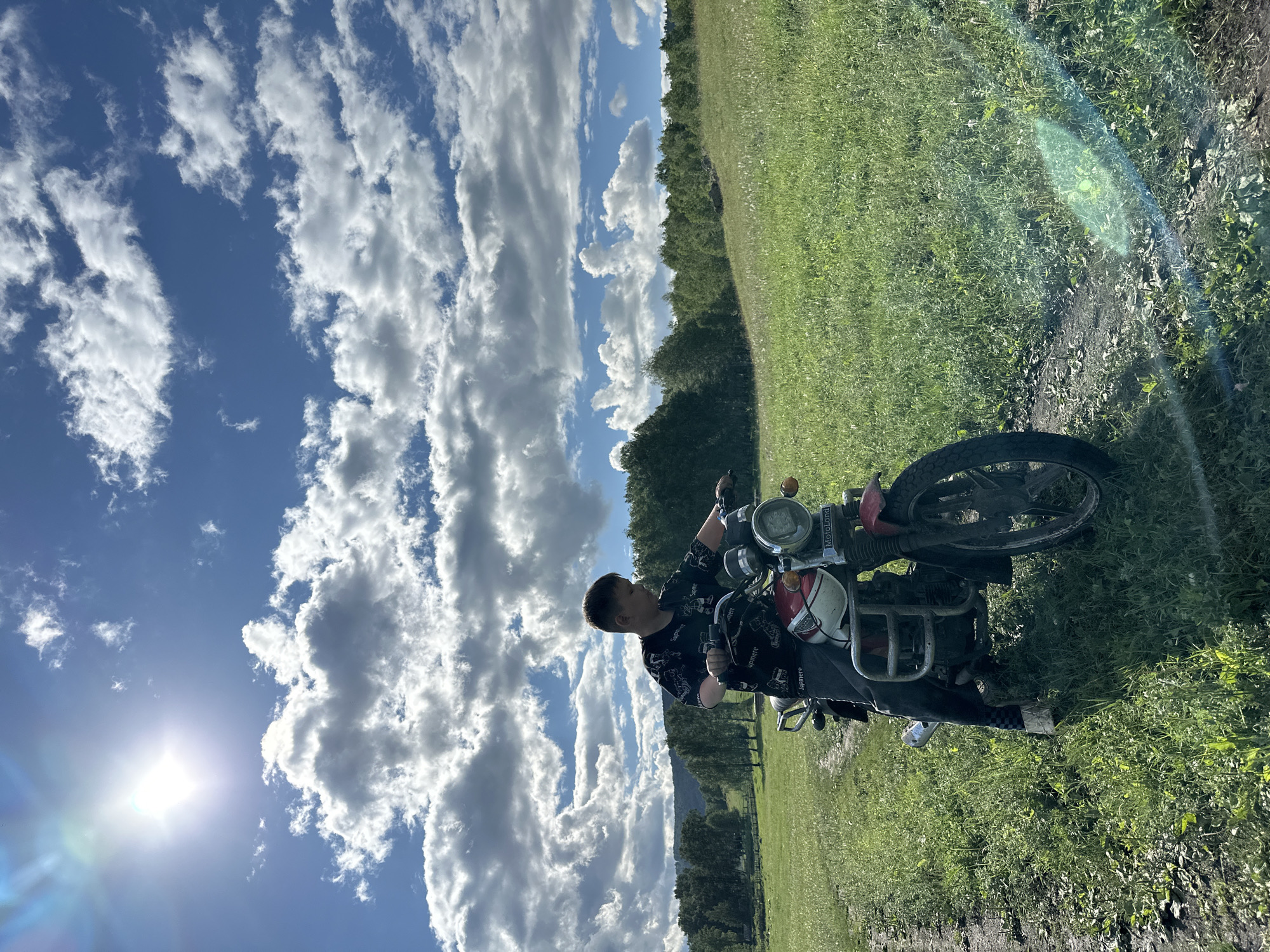 A young male is seated on a red and white motorcycle on a dirt path in a grassy field. The sky is blue with scattered white clouds, and the sun is visible in the upper left corner, creating lens flare. Trees form a treeline in the background, and hills are visible beyond the trees. The motorcycle has a round headlight, chrome handlebars, and a license plate that is unreadable. The male is wearing a dark t-shirt with a pattern and dark pants. The overall scene depicts an outdoor rural setting on a sunny day.