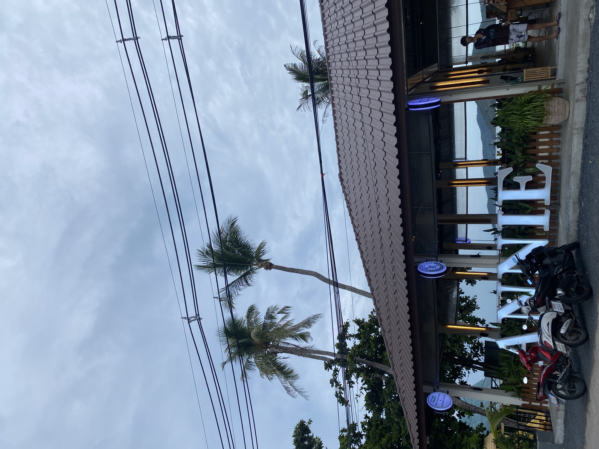 Captured in Baan Thong Krut, Thailand, the image presents an outdoor scene. The foreground features parked motorbikes in front of a building, with signage prominently displaying illuminated letters, apparently part of a business name. Palm trees are visible extending into the sky, interspersed with power lines. The sky is overcast. In the background, a body of water and a shoreline are faintly visible through the building's structure, suggesting a seaside location. A person is standing in the building's entrance.