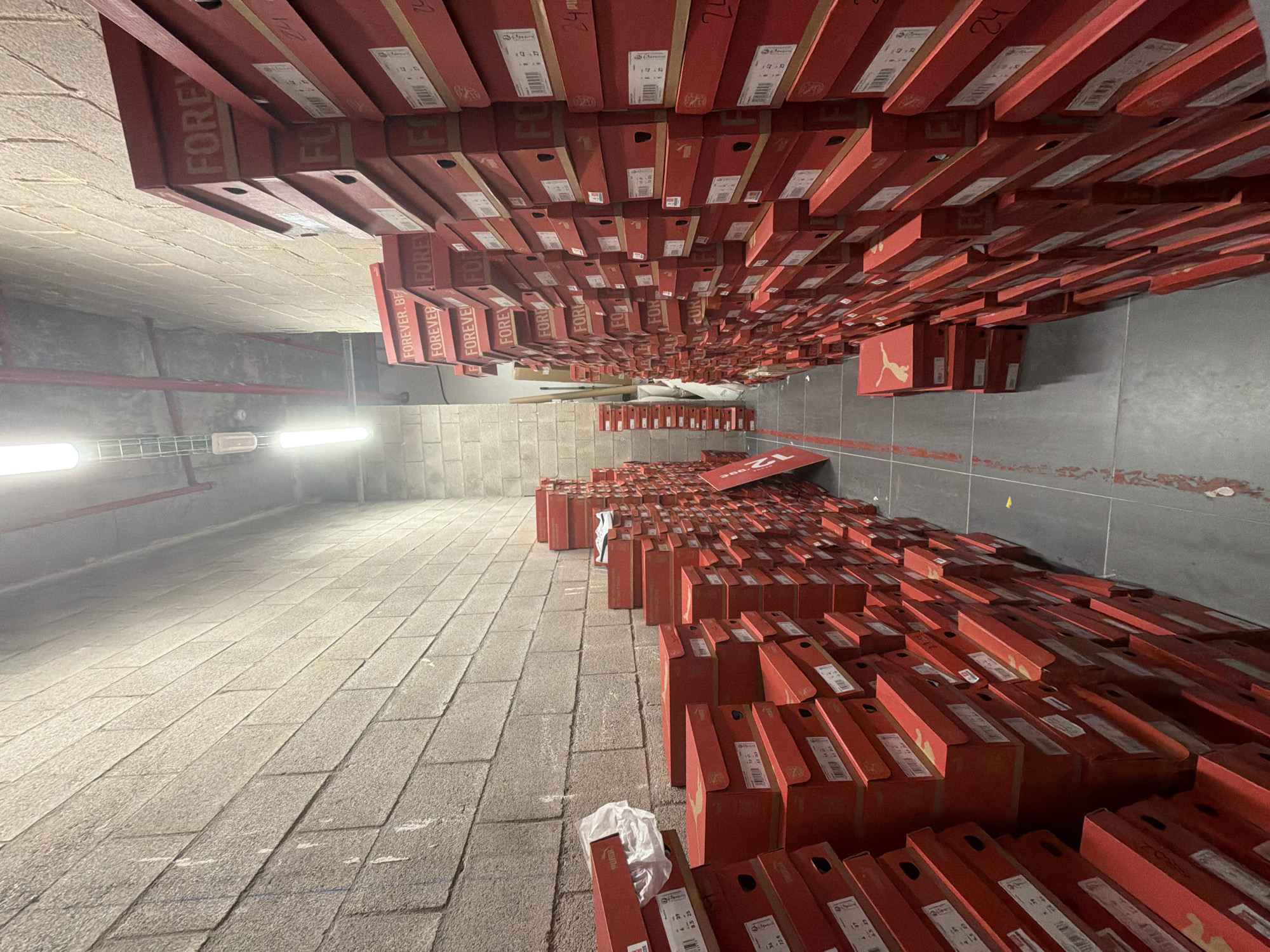 The image shows a long, narrow storage area with walls made of concrete blocks. The floor is a polished grey concrete. The space is filled with stacks of red shoe boxes, predominantly branded with "FOREVER." and "PUMA." The boxes are stacked high along both sides of the corridor, creating a sense of depth and abundance. Fluorescent lights are mounted on the ceiling, casting illumination on the scene. A single white sneaker is visible on top of one of the stacks. The image is a forensic, matter-of-fact depiction of a storage facility, likely a warehouse or stockroom, with no people present and no discernible actions or interactions taking place. The specific location in the city of Madrid, Spain cannot be determined from the image alone.