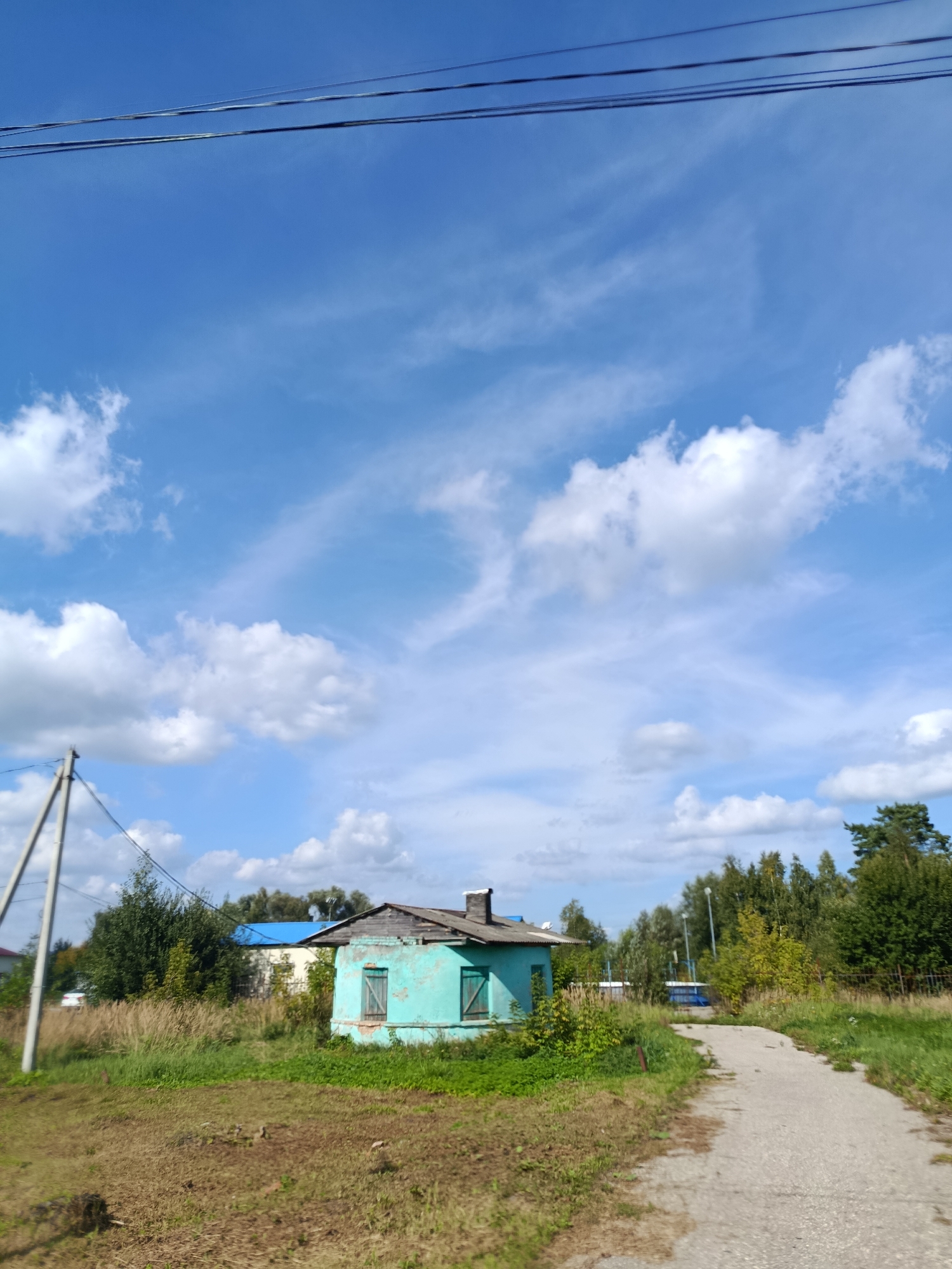 The image depicts a bright, partly cloudy day in a rural or semi-rural area. In the foreground, a dirt path curves to the right, bordered by dry grass and vegetation. To the left, a small, old, teal-colored house with wooden shutters and a damaged roof sits amongst overgrown greenery. Behind the house, a power pole with overhead wires is visible. Further in the background, a row of trees forms a treeline, with some buildings peeking through, one of which has a blue roof. A light-colored vehicle is partially visible to the left, and a blue object, possibly another vehicle or structure, is further back on the path. The sky is a vibrant blue with scattered white clouds. The scene appears to be in the city of Vereya, Russia.
