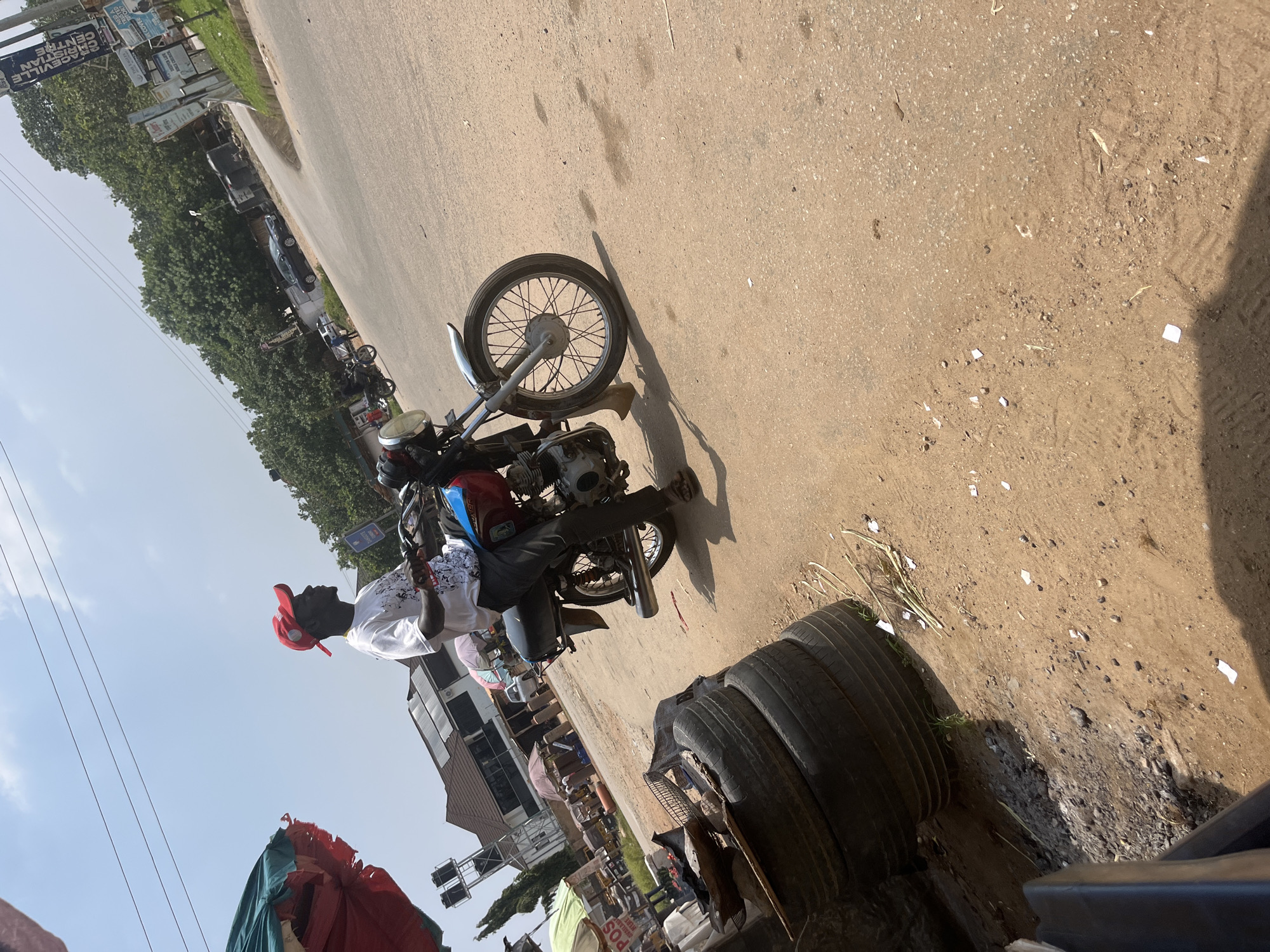 This media file depicts a street scene in Abuja, Nigeria, likely during the daytime. The primary subject is a person on a red and blue motorcycle, viewed from a slightly elevated angle. The person, wearing a white t-shirt with a black graphic design and a red baseball cap, is seated on the motorcycle. The motorcycle itself is a prominent object, with its spoked wheel, exhaust pipe, and engine clearly visible.
The setting is a paved road, appearing somewhat worn and dusty, with patches of dirt and debris scattered on the surface. To the left of the road, there is a line of trees, suggesting an urban or suburban environment. In the background, buildings and signage are visible, indicating commercial activity. One sign appears to read "GRACEVILLE CHRISTIAN CENTRE". Several other vehicles, including cars and other motorcycles, are parked or in motion along the road, contributing to a sense of a busy thoroughfare.
In the foreground, a stack of old tires is piled on the side of the road, adding a utilitarian element to the scene. The lighting suggests it is daytime, with shadows cast on the road, possibly indicating mid-morning or afternoon. The overall atmosphere seems casual and everyday, capturing a moment of transit or pause on a street in Nigeria.