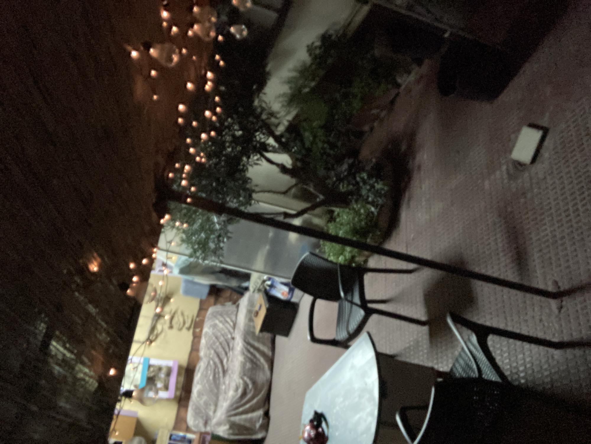 This is a nighttime outdoor scene, possibly a patio or courtyard, in Sabadell, Spain, characterized by low lighting and string lights overhead. In the foreground, a light-colored table with a dark base is positioned to the left, with two dark chairs nearby, one partially visible in the bottom left corner and another in the middle ground. The floor appears to be a textured, dark red surface.
Further back, a sofa covered with a patterned fabric is visible. To the right of the sofa is a dark, square end table with an object on top. Beyond this, there is a tree with dense foliage and a rough-textured wall or structure behind it. The overall environment suggests a casual outdoor seating area, possibly an extension of a residential or commercial space, illuminated by decorative string lights that are draped from a dark, possibly thatched, ceiling structure. There are no people visible in the image.