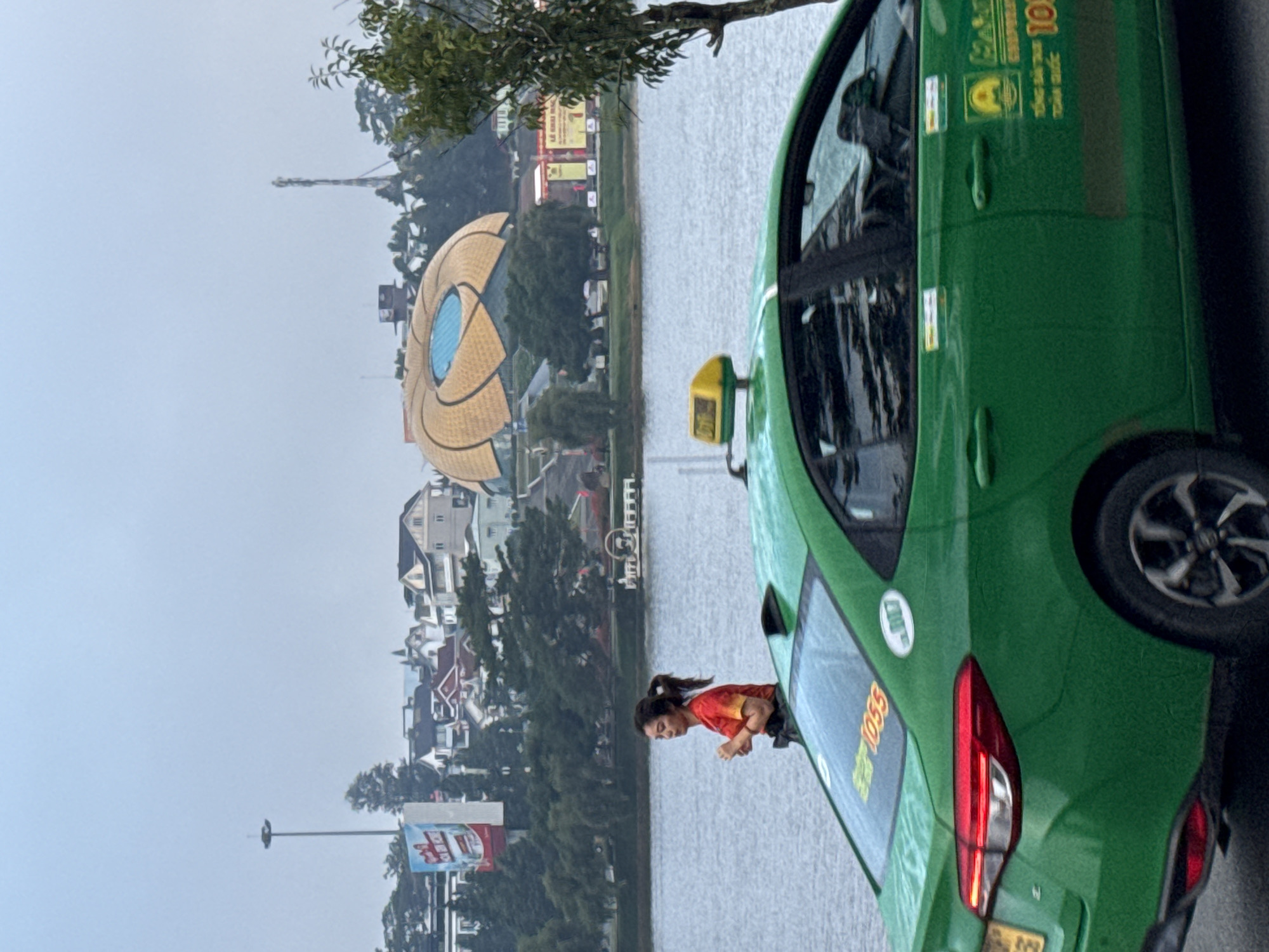 A green taxi is parked on a street next to a body of water. A young woman wearing a red and yellow shirt is standing on the sidewalk, looking out at the water. In the background, there are buildings and trees. The taxi has a yellow sign on its roof and the number "1055" is visible on the rear window. The scene appears to be in Da Lat, Vietnam, a city known for its lakes and French colonial architecture. The overall atmosphere is calm and serene, with the water reflecting the overcast sky.
