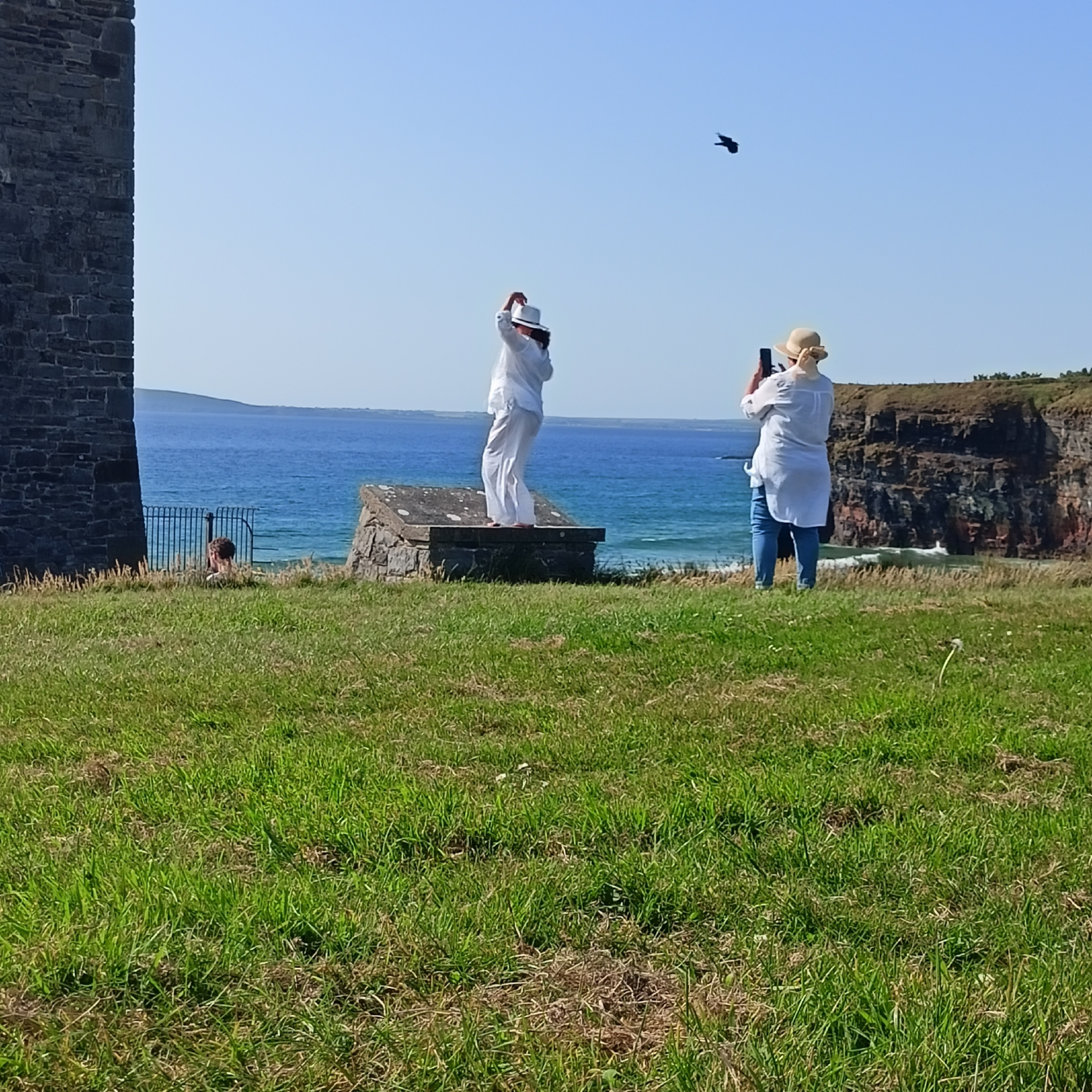 The image depicts a scenic coastal view in Ballybunnion, Ireland. In the foreground, a grassy hill slopes down towards the ocean, with a rough stone structure visible on the left, likely part of an old building or ruin. Two women are the primary human subjects. The woman on the left is standing on a stone pedestal, wearing a white outfit and a white hat, with her arms raised as if posing or gesturing. She appears to be looking towards a bird in flight. The woman on the right is further back, also wearing white and a straw hat, and is holding a smartphone to take a photograph. She is looking towards the right, in the direction of a dramatic cliff face that dominates the background. The ocean stretches out beyond the cliff, with waves crashing against its base. The sky is clear and blue, with a single dark bird visible in flight between the two women. A metal fence runs along the edge of the grassy area, separating it from the cliff and ocean. Another person is partially visible behind the fence, near the stone structure on the left. The overall atmosphere is one of a bright, sunny day at a popular tourist location.