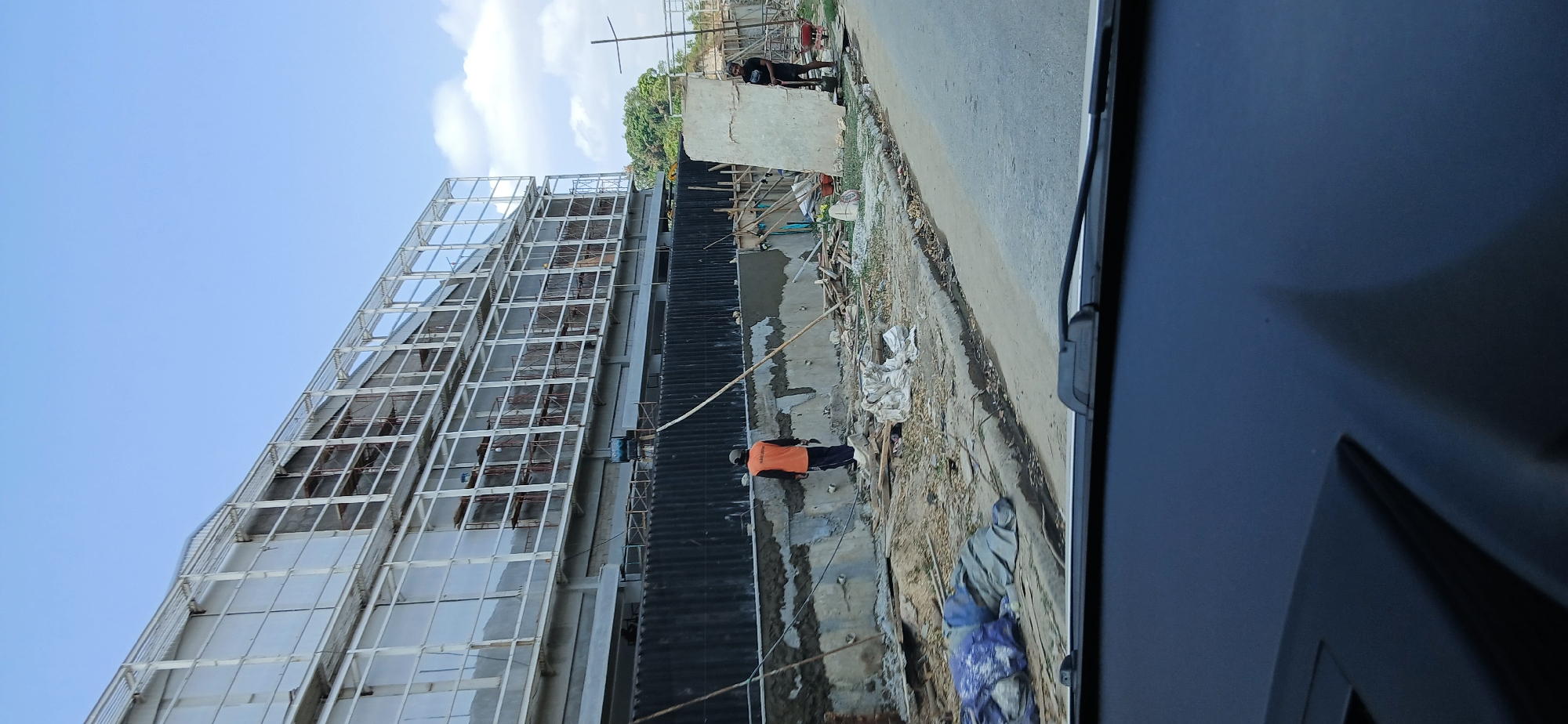 The image depicts a construction site with a large building under construction in Samarinda, Indonesia. Two construction workers are visible. One worker, wearing an orange vest, stands near a partially constructed wall. Another worker, wearing a dark t-shirt, is using a shovel near a pile of material. The building features a white metal framework and large, rectangular panels. The ground is uneven and covered with debris, including wood scraps and tarpaulins. The sky is blue with a few clouds. The foreground shows the dashboard of a vehicle.