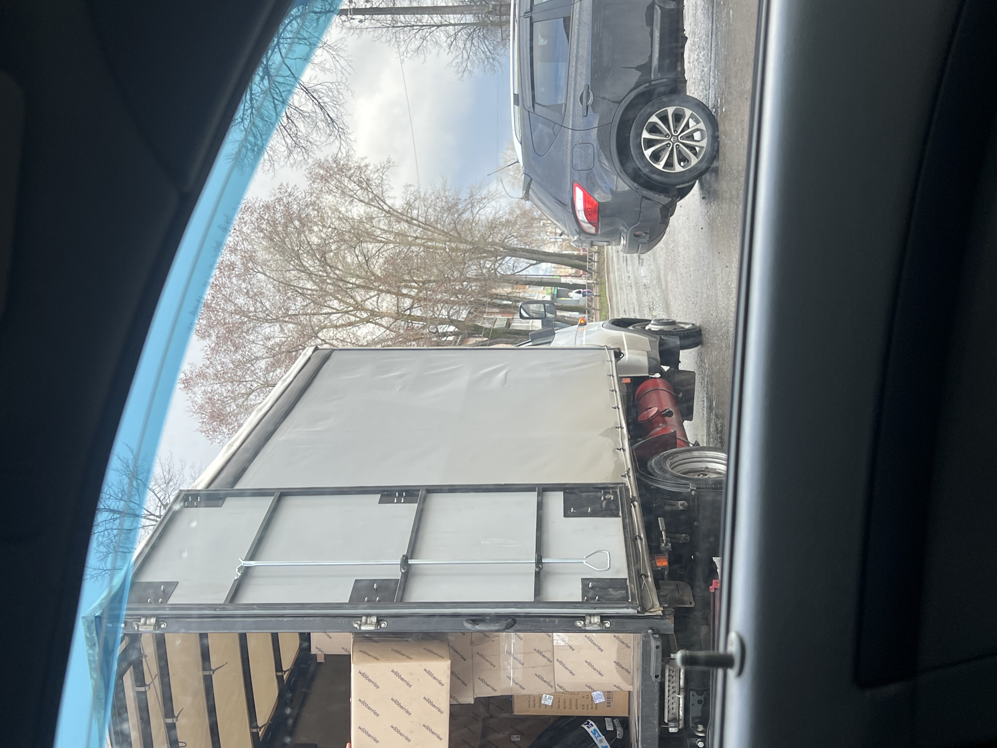 The photograph, taken from inside a vehicle, shows a view of a truck and a dark SUV outside the vehicle. The back of the truck is open, revealing boxes inside, labeled "wildberries". The truck has a gray tarp covering part of its cargo area. To the side, there is a dark SUV, with its rear tire visible. Outside the vehicles, bare trees suggest a city street environment. The photo location is Chaikovsky, Russia, as identified by the wildberries branding on the boxes, a popular Russian online retailer.