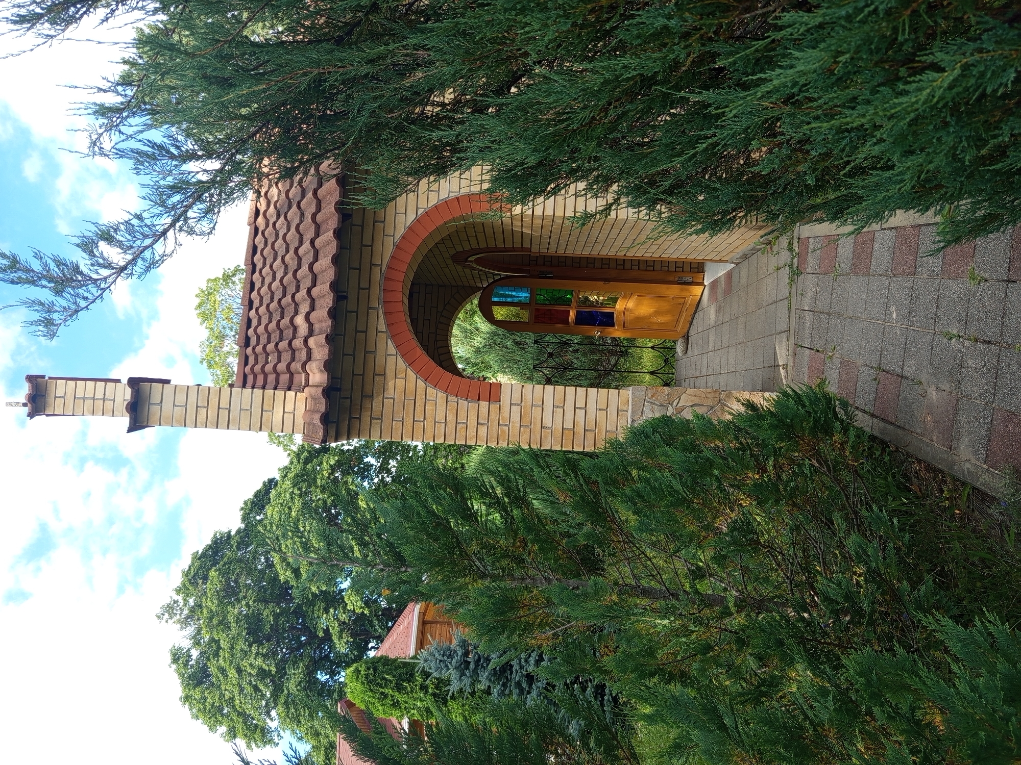 An outdoor, eye-level, full shot shows a path leading to an arched entryway of a brick building. The entryway has a wooden door with stained glass panels and an open archway above it, revealing more greenery beyond. The building has a tiled roof and a brick chimney. The path is paved with square tiles of gray and red. Lush green trees and bushes surround the building, with a clear blue sky with white clouds overhead. The scene is set in the city of Giaginskaya, Russia.