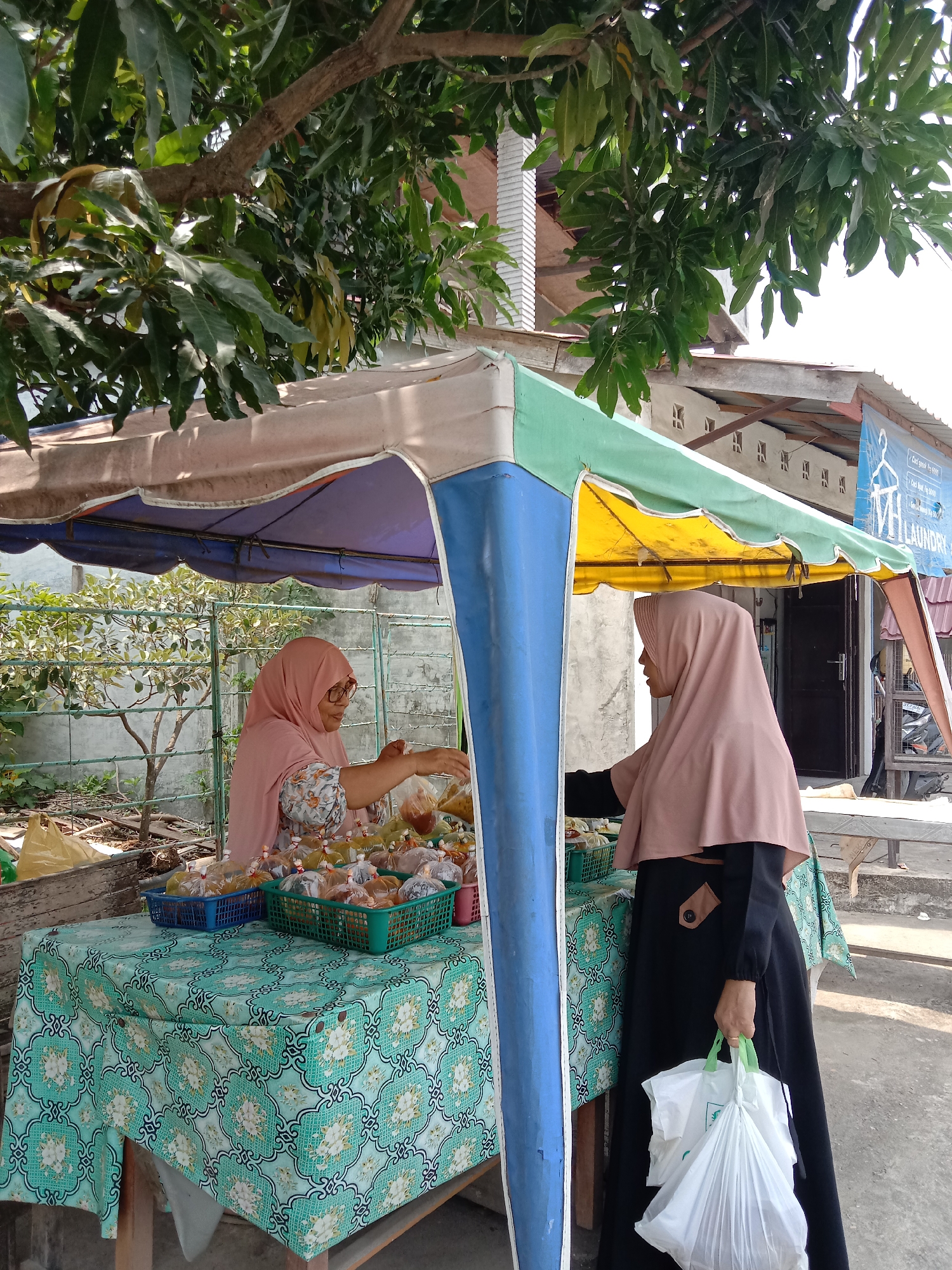 The image captures a scene at an outdoor food stall in Lhokseumawe, Indonesia. A woman wearing a peach-colored hijab and floral patterned blouse is attending to a customer. The customer, also wearing a hijab (peach-colored), is dressed in a black abaya and holding two white plastic shopping bags with green handles. The food stall is sheltered by a canopy of multi-colored fabric (blue, yellow, green, and beige). The stall's table is covered with a patterned tablecloth in shades of green and turquoise. Baskets of pre-packaged food items are arranged on the table. The setting appears to be a residential neighborhood with trees overhead and a building with a "Laundry" sign visible in the background.