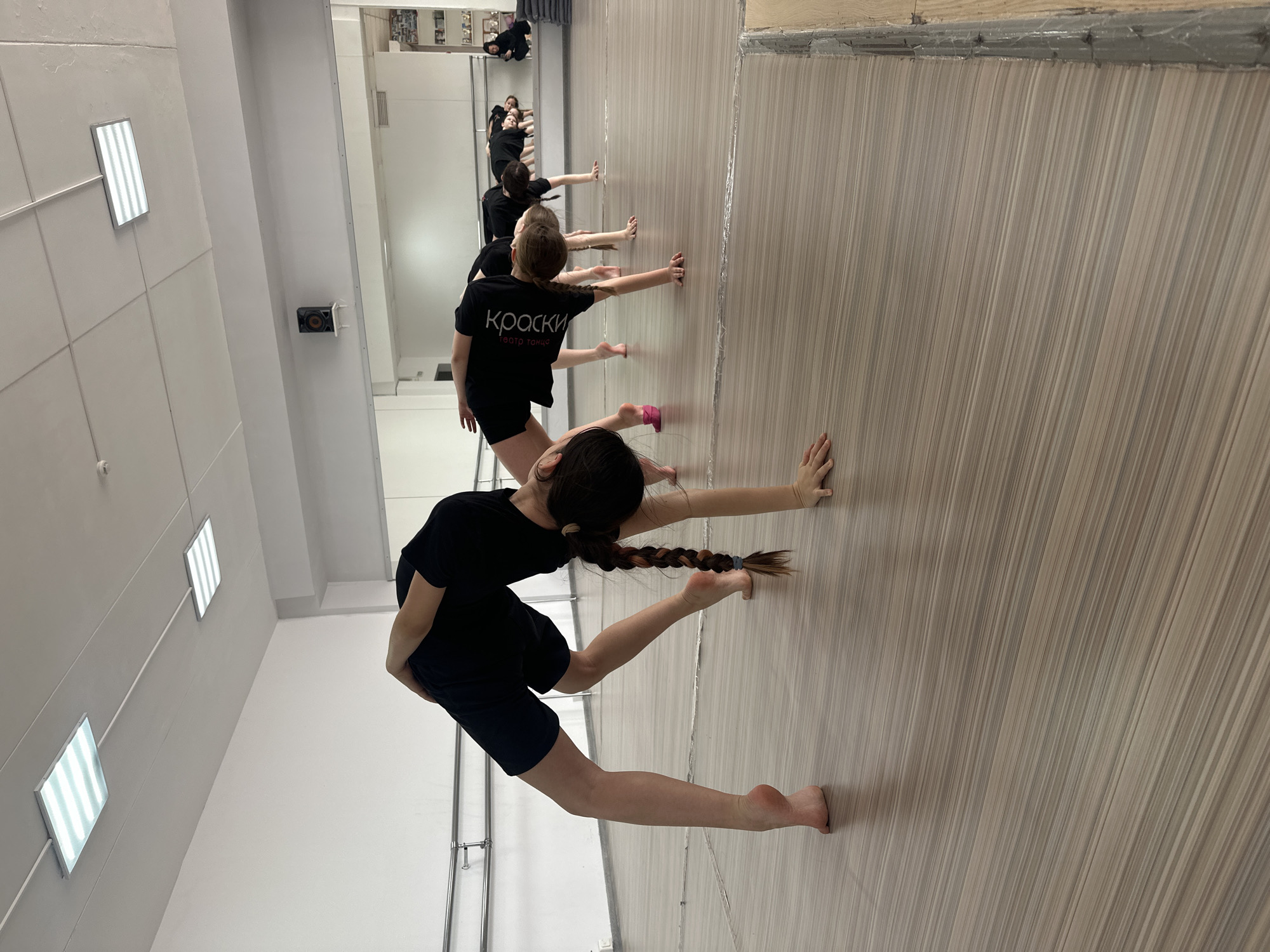 In a brightly lit studio in Votkinsk, Russia, a dance class is in progress. Several young girls wearing black t-shirts and shorts are practicing a ballet pose, pressing their hands against a striped wooden wall. The girls are aligned in a row, with the first dancer in the foreground executing a more advanced variation of the pose. A mirror on the adjacent wall reflects the dancers and the studio space. The ceiling features rectangular LED panel lights, and a ventilation system is visible on the wall.