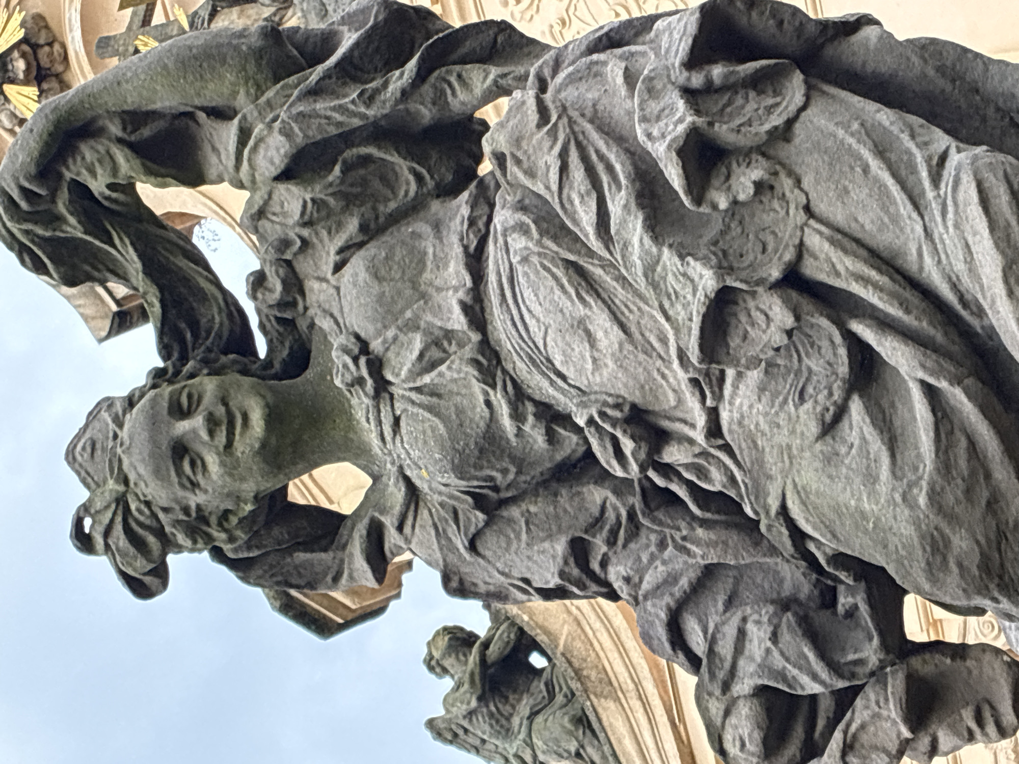 The image shows a stone statue of a female figure with flowing drapery, positioned outdoors against a cloudy sky. The statue is weathered and covered in moss, indicating it is an old monument. The figure's face is smiling, and her hair is adorned with a ribbon-like headdress. In the background, another, smaller stone statue of an angel can be seen, also weathered and partially obscured. The architectural style suggests a Baroque or Rococo period. This statue is likely part of the Kuks Hospital complex in Kuks, Czechia, which is known for its collection of Baroque sculptures by Matthias Braun.