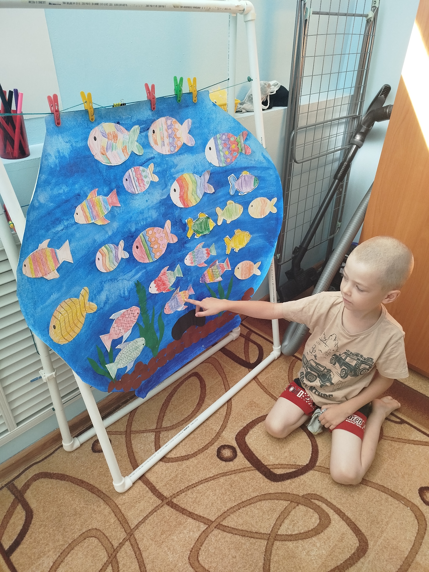 A young, light-skinned boy with a shaved head and wearing a light brown t-shirt with a graphic design and red shorts kneels on a beige carpet with brown swirling patterns. He is pointing his left index finger towards a large, blue, cutout resembling an aquarium, which is adorned with numerous colorful, paper cut-out fish. The aquarium cutout is attached to a white PVC pipe frame and suspended by clothespins. To the left of the aquarium cutout, a red cup holds several markers. Behind the aquarium, a metal drying rack and a vacuum cleaner are visible. A wooden door is partially visible on the right side of the frame. The scene appears to be indoors, possibly in a room in Zavodoukovsk, Russia.