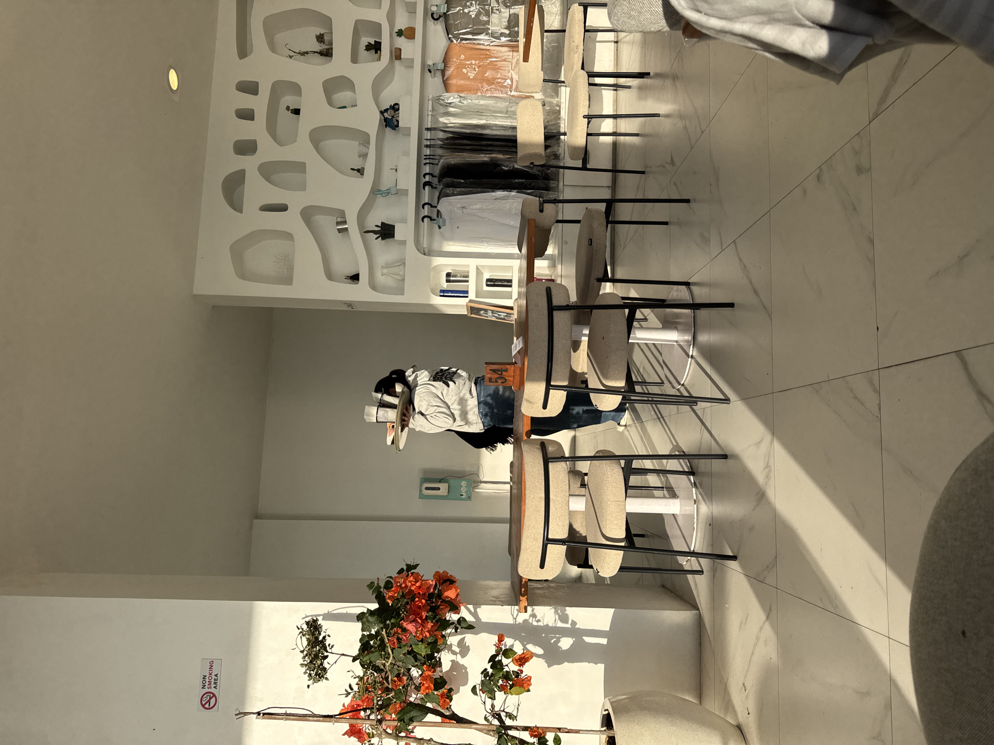 Here is a description of the image.
The image shows the interior of a retail space, possibly a clothing or accessory boutique in Kedisan, Indonesia. The space has a modern and clean aesthetic with white walls and light-colored tile flooring.
To the left, a wall display with organically shaped recessed shelves holds various decorative items, including small plants, vases, and sculptural objects. Clothing items hang on racks along the back wall.
A central area features a series of tables with tall, upholstered stools. A person in casual wear and a wide-brimmed hat stands behind one of the tables, interacting with it.
In the lower left corner, there is a potted plant with vibrant orange flowers. A "Non Smoking Area" sign is visible on the wall near the plant.
The room is brightly lit with natural light streaming in from the right side of the frame.