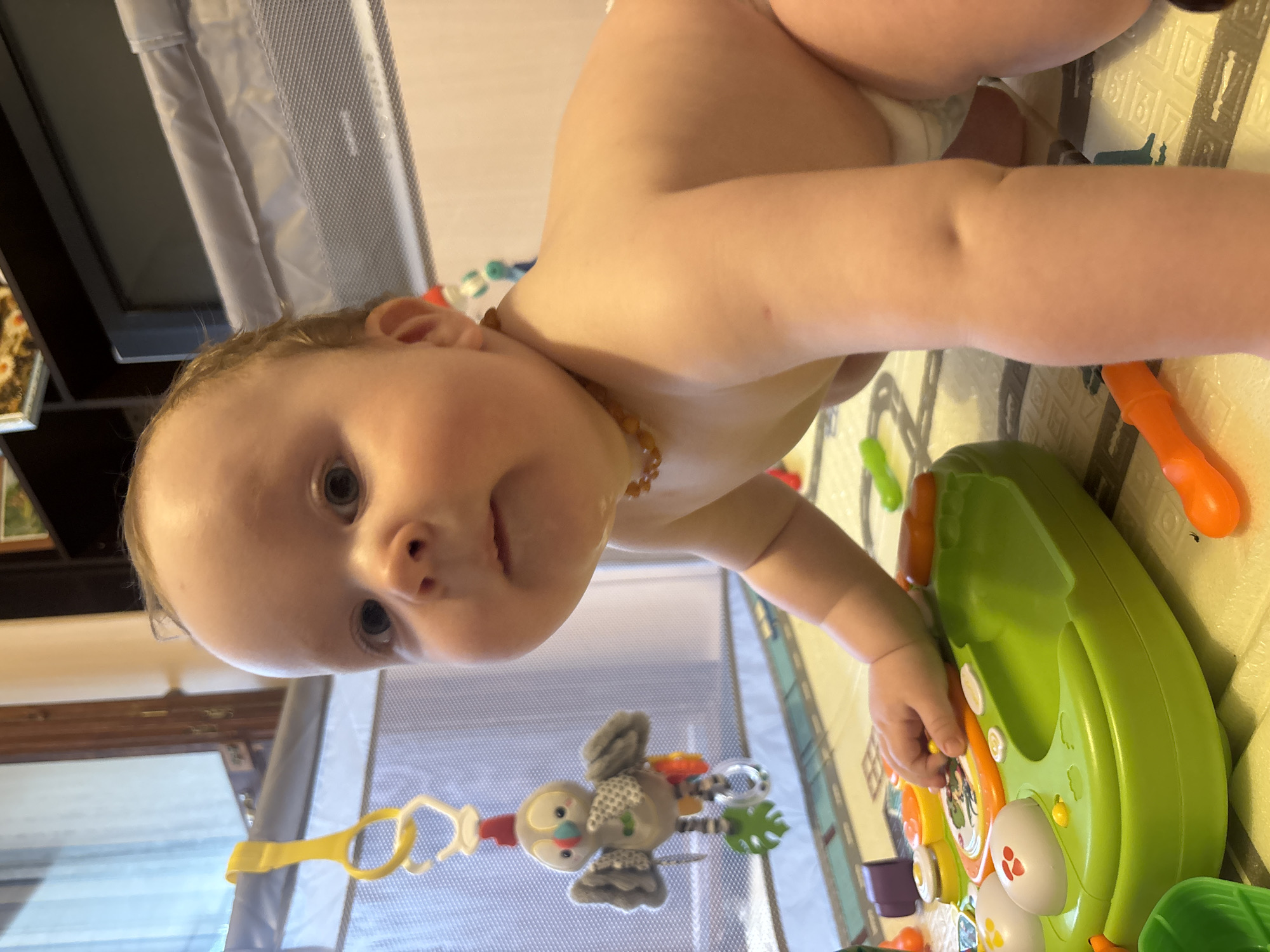 A baby wearing a diaper and an amber necklace is in a playpen with a padded play mat. The baby is positioned on their stomach, reaching for a green and orange toy. A toy mobile with a grey bird hangs above the playpen. The location is inferred from metadata, but there are no recognizable landmarks visible in the image to corroborate the city of Марковский, Russia. The environment appears to be indoors, with a playpen and other furniture visible in the background.