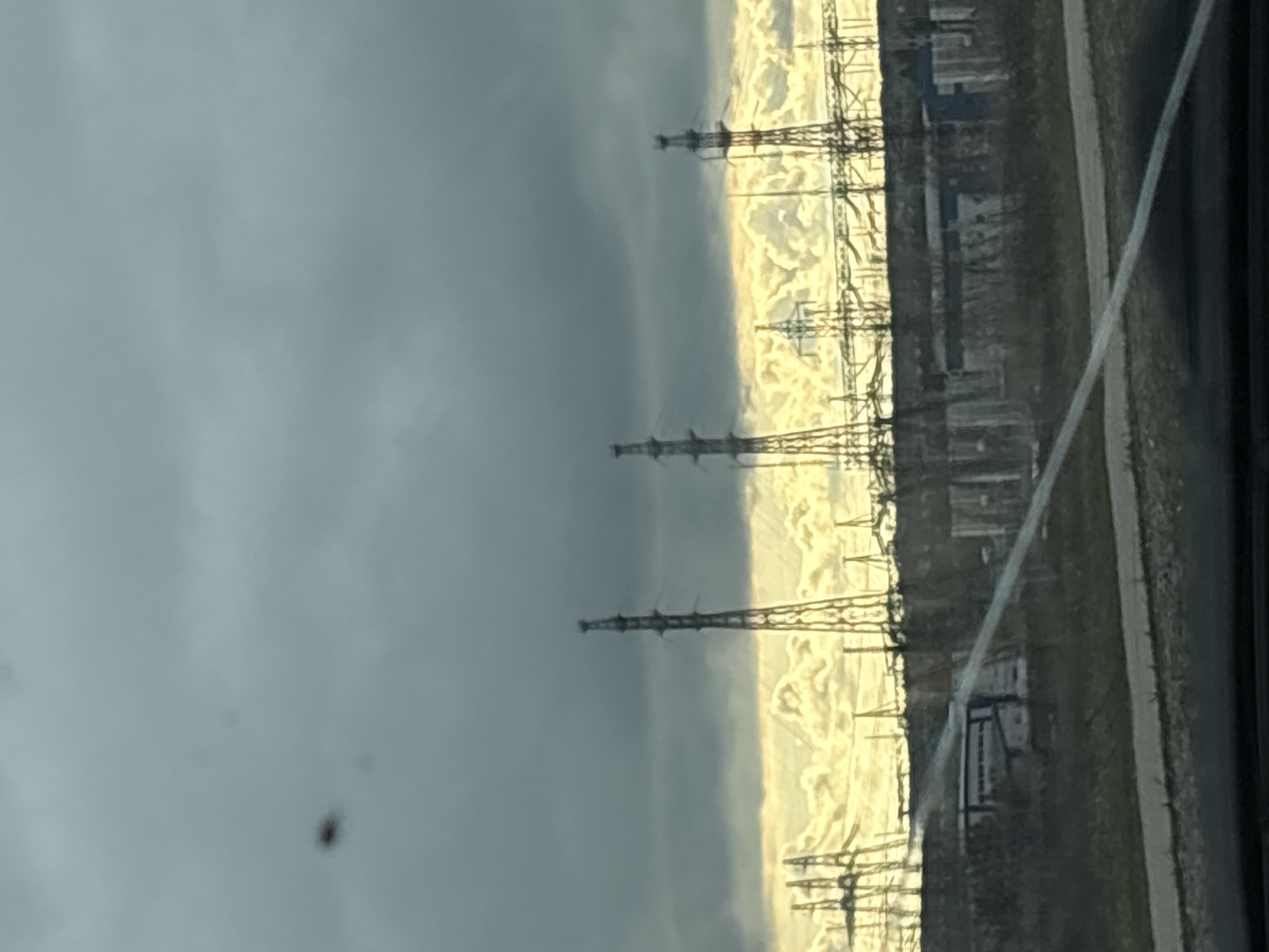 The image, rotated 90 degrees clockwise, captures a dramatic, overcast landscape featuring power infrastructure under a striking sky. The upper portion of the frame is dominated by a dark, heavy grey sky, suggesting stormy weather or a late hour. Below this, a bright, almost golden band of light stretches across the horizon, appearing to be the sun breaking through low clouds or reflecting off them.
Silhouetted against this luminous horizon are multiple tall power transmission towers, easily identifiable by their lattice-like structures. To their right, extending further along the horizon, is a power substation with various dark, industrial-looking buildings and equipment.
The ground area, below the horizon, is dark and muted, possibly covered in sparse vegetation. A lighter-colored, narrow path or road runs horizontally across this lower section of the image. A thin, diagonal, bright line is visible in the bottom right, likely a reflection or mark on the window or lens through which the photo was taken. A small, blurry, dark speck, possibly a bug or dirt, is also present in the dark sky area.
The scene conveys a sense of a powerful, possibly solemn, atmosphere due to the dramatic contrast between the dark sky and the glowing horizon. The time of day appears to be late afternoon or early evening. No visible text is present in the image.