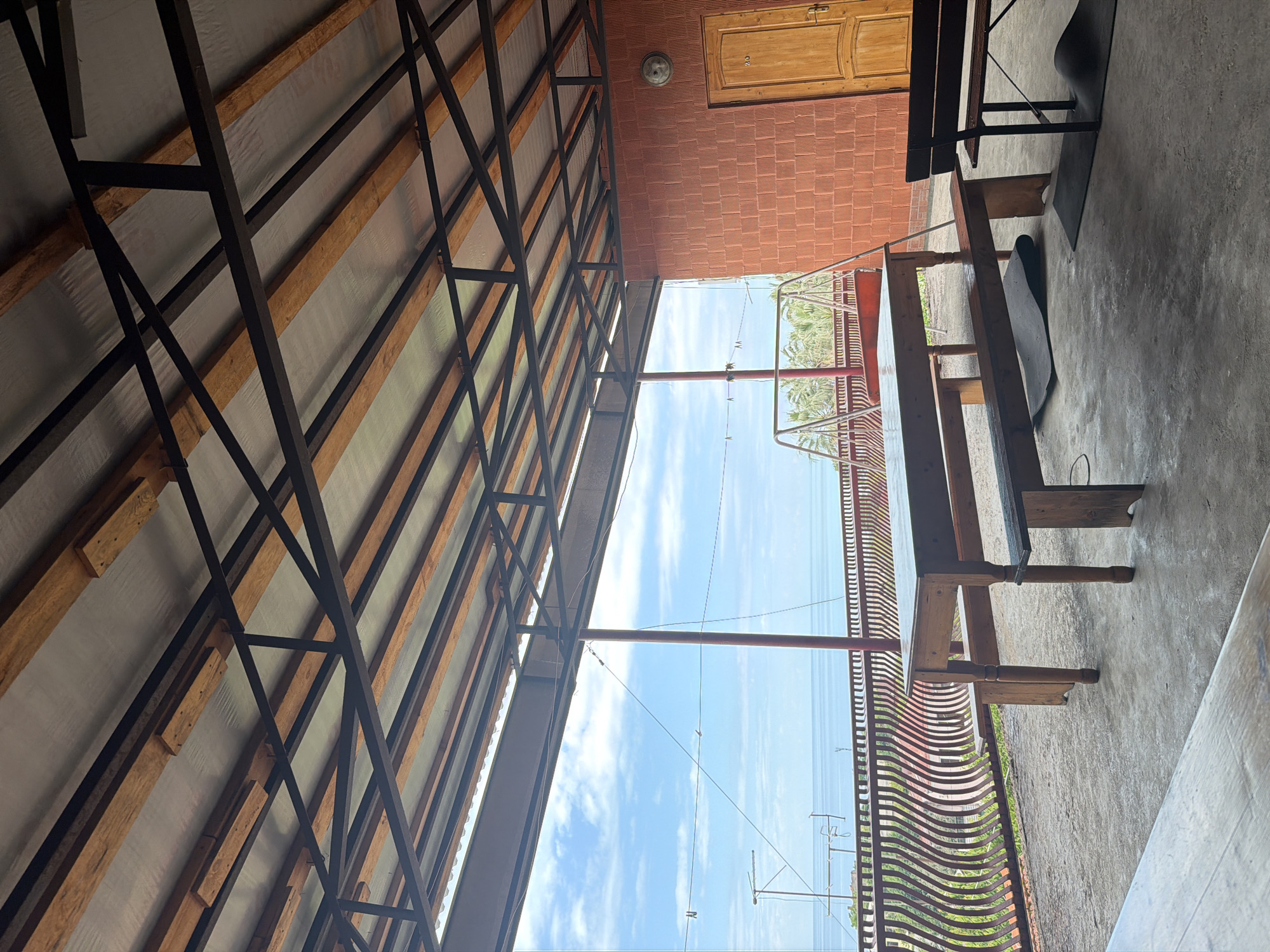 The image shows an outdoor seating area with a covered roof supported by a metal and wood structure. In the foreground, there is a concrete floor with a wooden table and two benches. To the right, there is a wall made of textured orange bricks with a wooden door. In the background, a metal railing with a wavy design overlooks a body of water and a blue sky with scattered clouds. There is also a swing set visible behind the railing. The scene appears to be a dining or relaxation area at a coastal location. There are no people or animals present in the image.