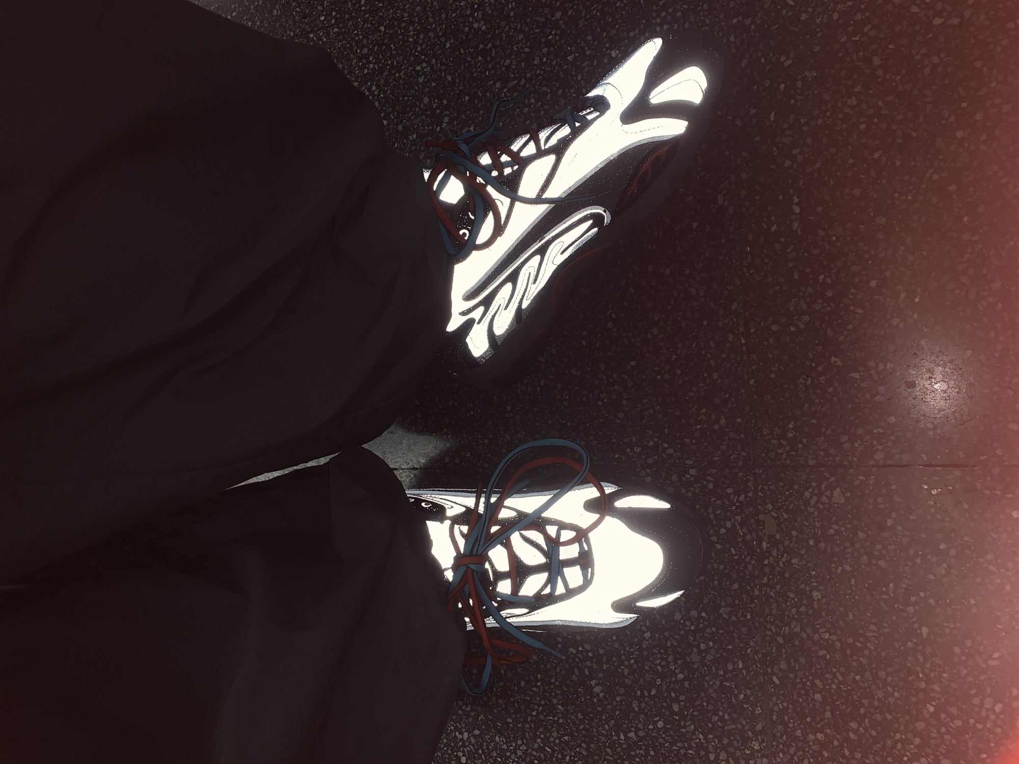 The photograph shows a top-down view of a person's feet, clad in dark pants and sneakers, standing on a dark-colored speckled surface that appears to be a city sidewalk or pavement. The sneakers are lit intensely by reflected light, possibly a flash, accentuating their design. The laces of the sneakers are blue and orange. The ground appears to be made of small stones. The image has a vintage or slightly faded quality. The location is not discernible as Barcelona, Spain.