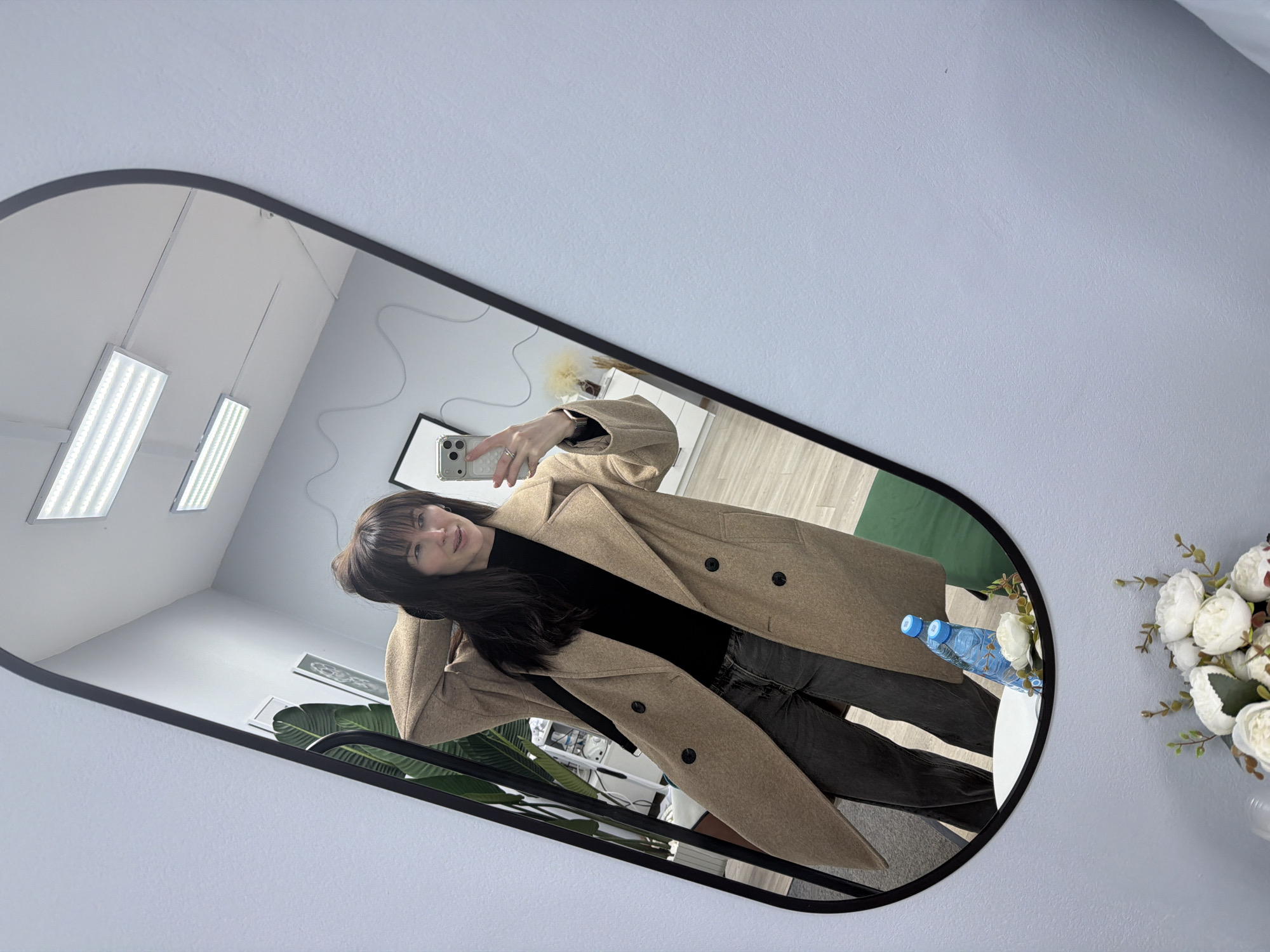 The image depicts a mirror selfie of a woman taken in an indoor setting, likely in Perm, Russia. The woman, appearing to be in her 20s or 30s, has long dark hair with bangs and a subtle, pleasant smile. She is wearing a black top, dark jeans, and a long, light brown or camel-colored double-breasted coat. She holds a smartphone in her right hand, visible within the mirror's reflection.
The mirror itself has an irregular, rounded rectangular shape with a black frame. Inside the reflection, the background reveals a modern and well-lit room with white walls and a light-colored wooden floor. Two rectangular fluorescent light fixtures are visible on the ceiling, suggesting a commercial space, office, or studio. Other objects reflected include framed pictures on the wall, a large green leafy plant, and a white table with two blue plastic water bottles.
Outside the mirror's frame, the surrounding wall is painted a light blue-grey. To the right, a white vase holds an artificial bouquet of white flowers, possibly peonies or roses, with green foliage, adding a decorative element. The overall scene conveys a clean, contemporary interior. No visible text is present.