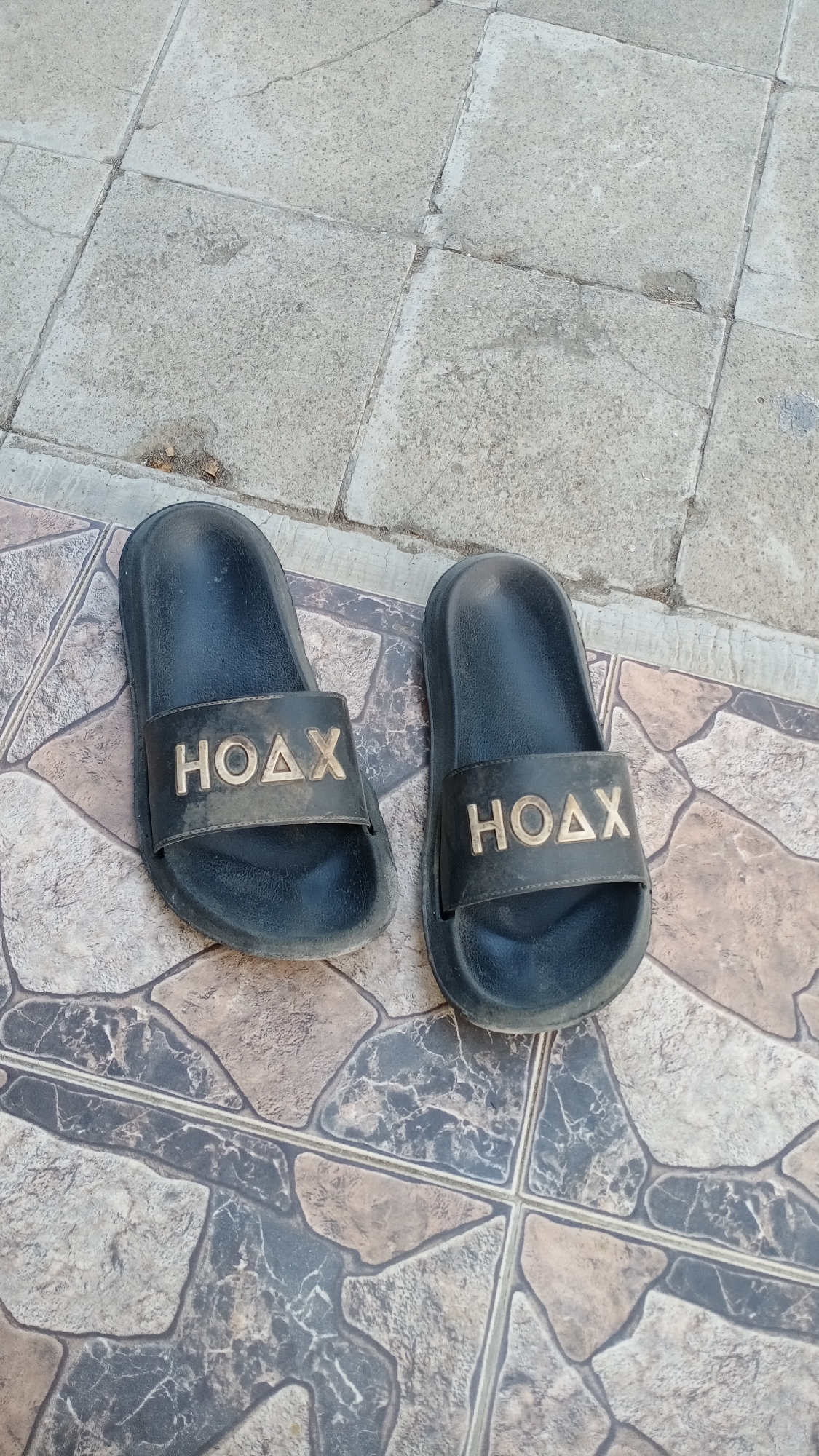 A pair of black "HOAX" brand slides are positioned on a tiled outdoor surface. The slides are slightly dirty and show wear. The left slide is closer to the camera and angled slightly outward, while the right slide is positioned further away and parallel to the left. The floor is composed of large, irregularly shaped tiles with a marbled pattern in shades of brown, beige, and gray. In the upper portion of the image, the surface changes to large, square, gray concrete tiles with visible cracks and some debris. The overall impression is of an outdoor pathway or patio. The image does not provide any specific information about the city of Mertan, Indonesia, or any people present.