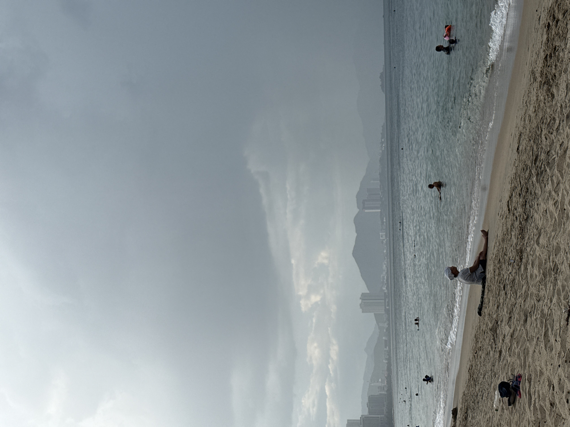 The image shows a cloudy day at a beach in Nha Trang, Vietnam. In the foreground, a person sits on the sand, facing the ocean. The person is wearing a light-colored shirt and dark shorts, with a white head covering. Several pairs of flip-flops and a water bottle are on the sand near the person. The ocean has small waves, and several people are swimming or playing in the water. In the background, a city skyline with tall buildings and mountains can be seen. The sky is filled with heavy, gray clouds, suggesting an overcast or potentially stormy day.