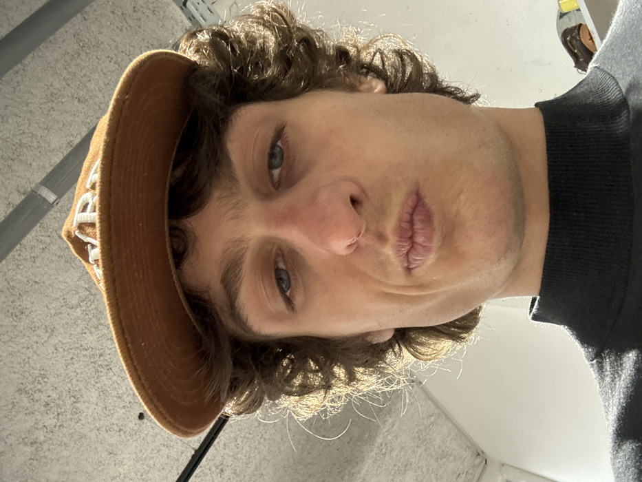 A person's face dominates the frame, taken from a low angle. The individual has light skin, blue eyes, and brown, curly hair. A brown baseball cap is partially visible on their head, with white lettering. Their lips are pursed slightly. They are wearing a dark-colored, high-necked sweater. The background consists of a ceiling with some visible fixtures and a wall. The image's perspective gives a slightly distorted view of the subject's features.